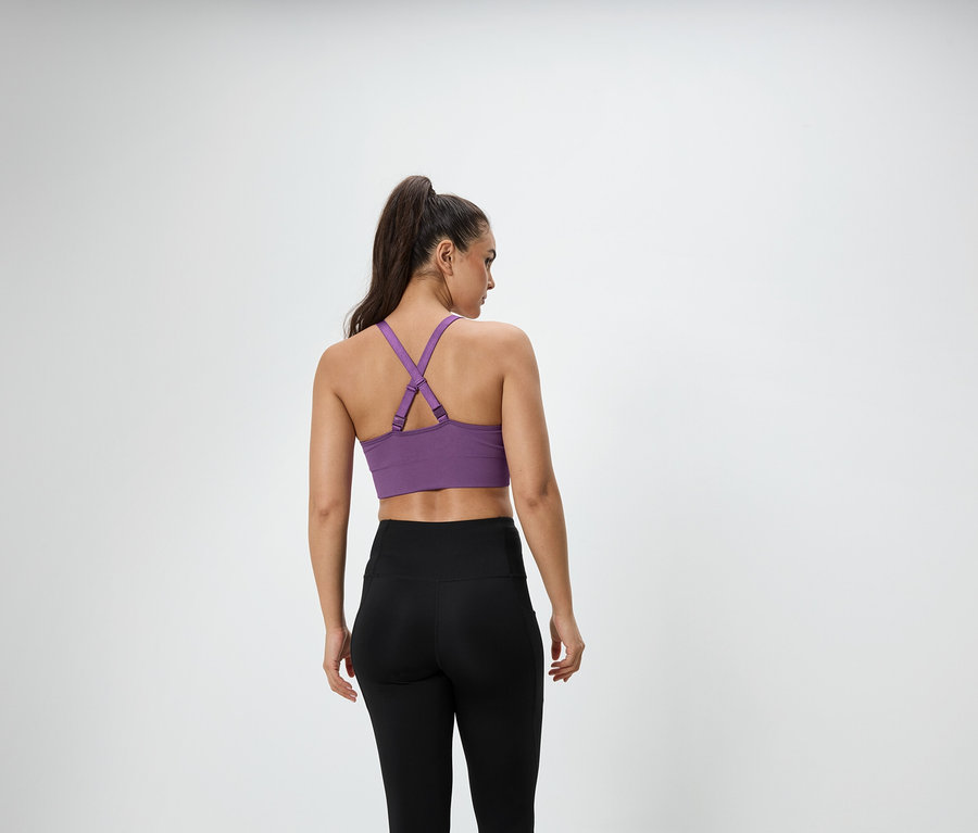 Kobieta z brązowymi włosami związanymi w kucyk stoi tyłem do kamery, ma na sobie fioletowy stanik sportowy i czarne legginsy.