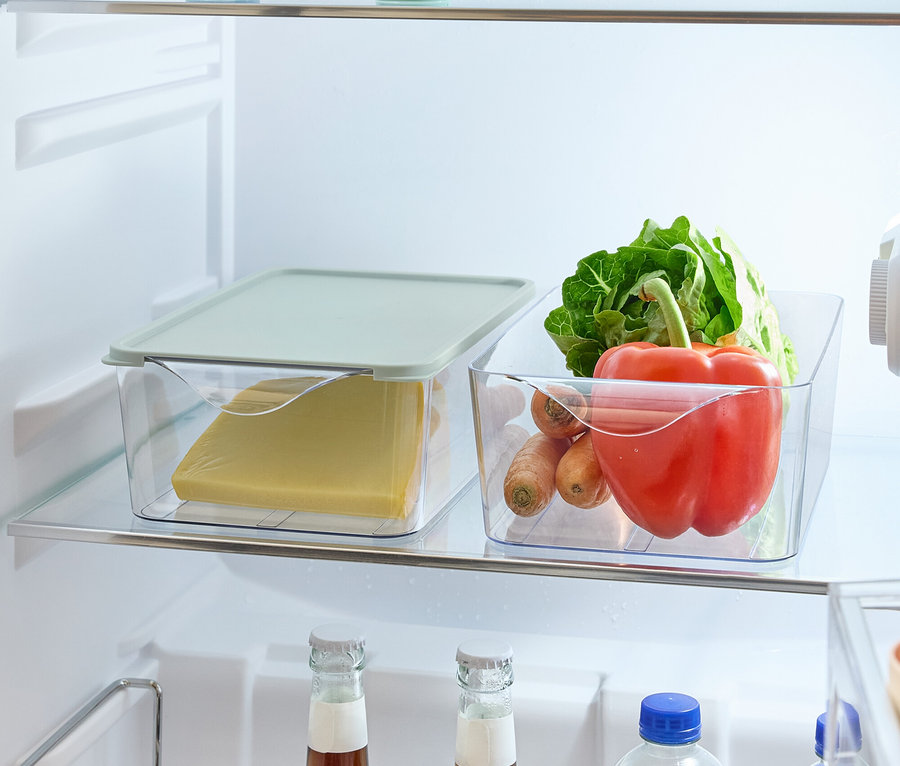 W lodówce na półkach znajdują się produkty spożywcze: jabłko i cytryna w okrągłych pojemnikach, ser w plastikowym pojemniku z pokrywką, warzywa (sałata, papryka, marchew) w plastikowym pojemniku oraz butelki z napojami w uchwycie.