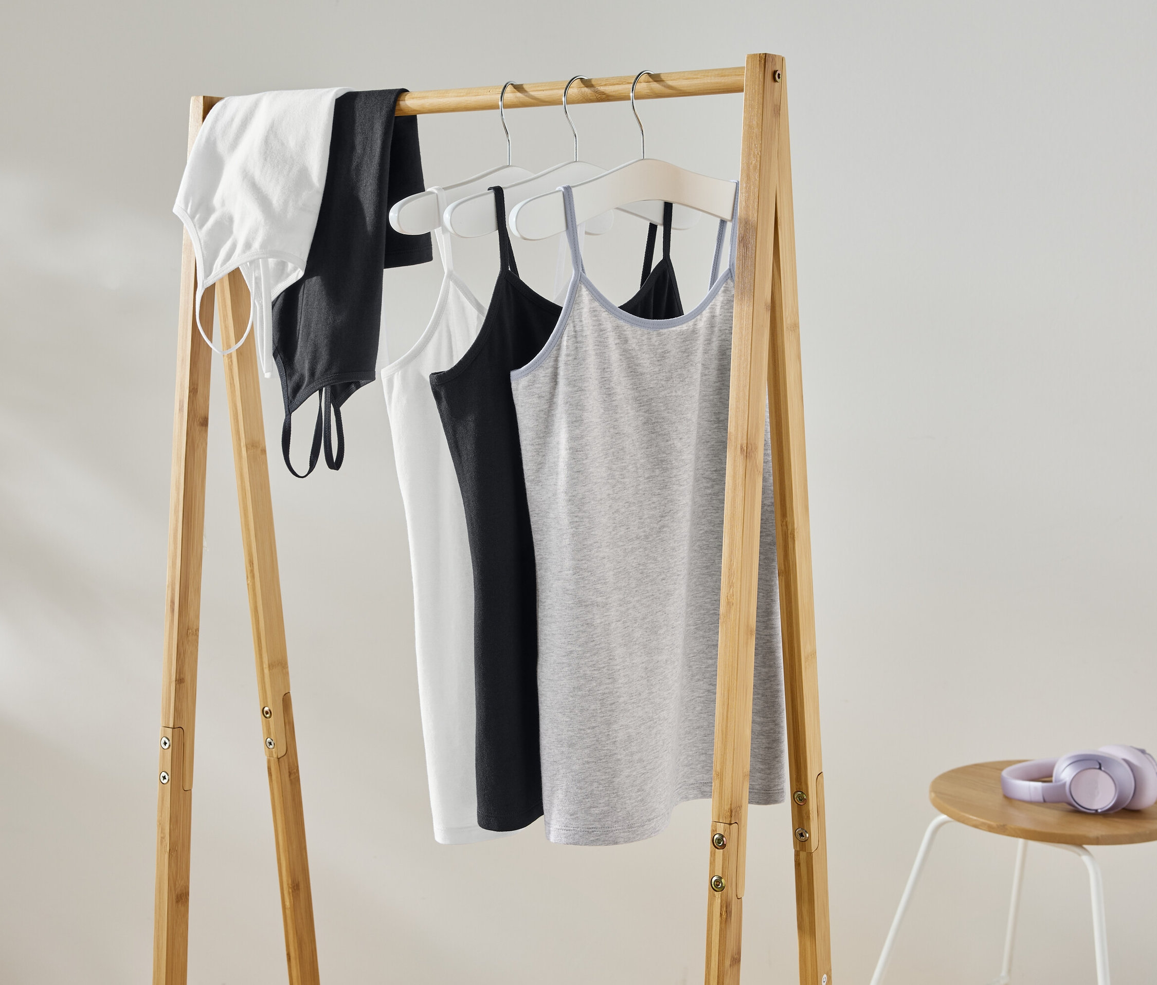 Na drewnianym wieszaku wiszą biały top, czarny top, biały podkoszulek, czarny podkoszulek i szary podkoszulek. Na stołku obok leżą fioletowe słuchawki.