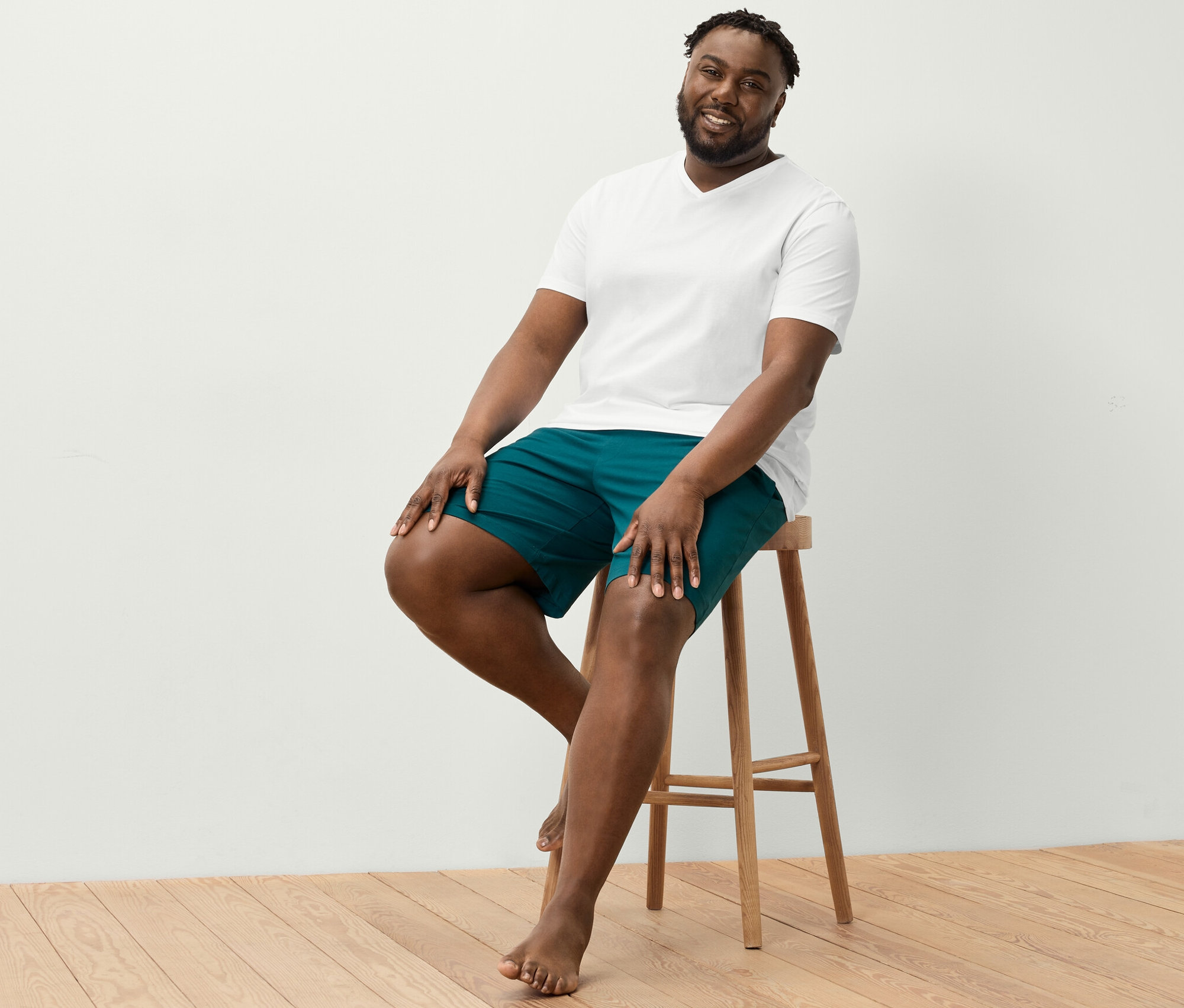 Mężczyzna siedzi na stołku ubrany w białą koszulkę z dekoltem w serek i krótkie spodenki rekreacyjne.