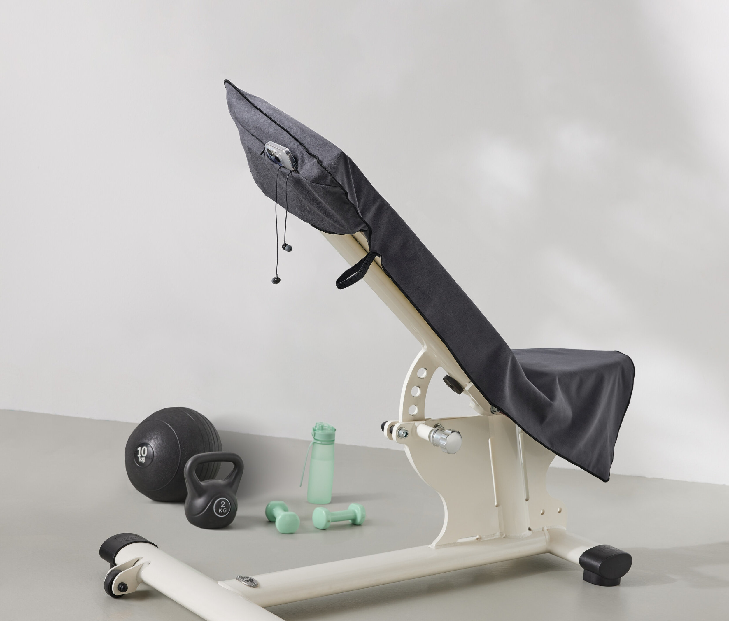 Ławka do ćwiczeń przykryta jest szarą tkaniną. Obok ławki leżą piłka lekarska, kettlebell, butelka z wodą i hantle.