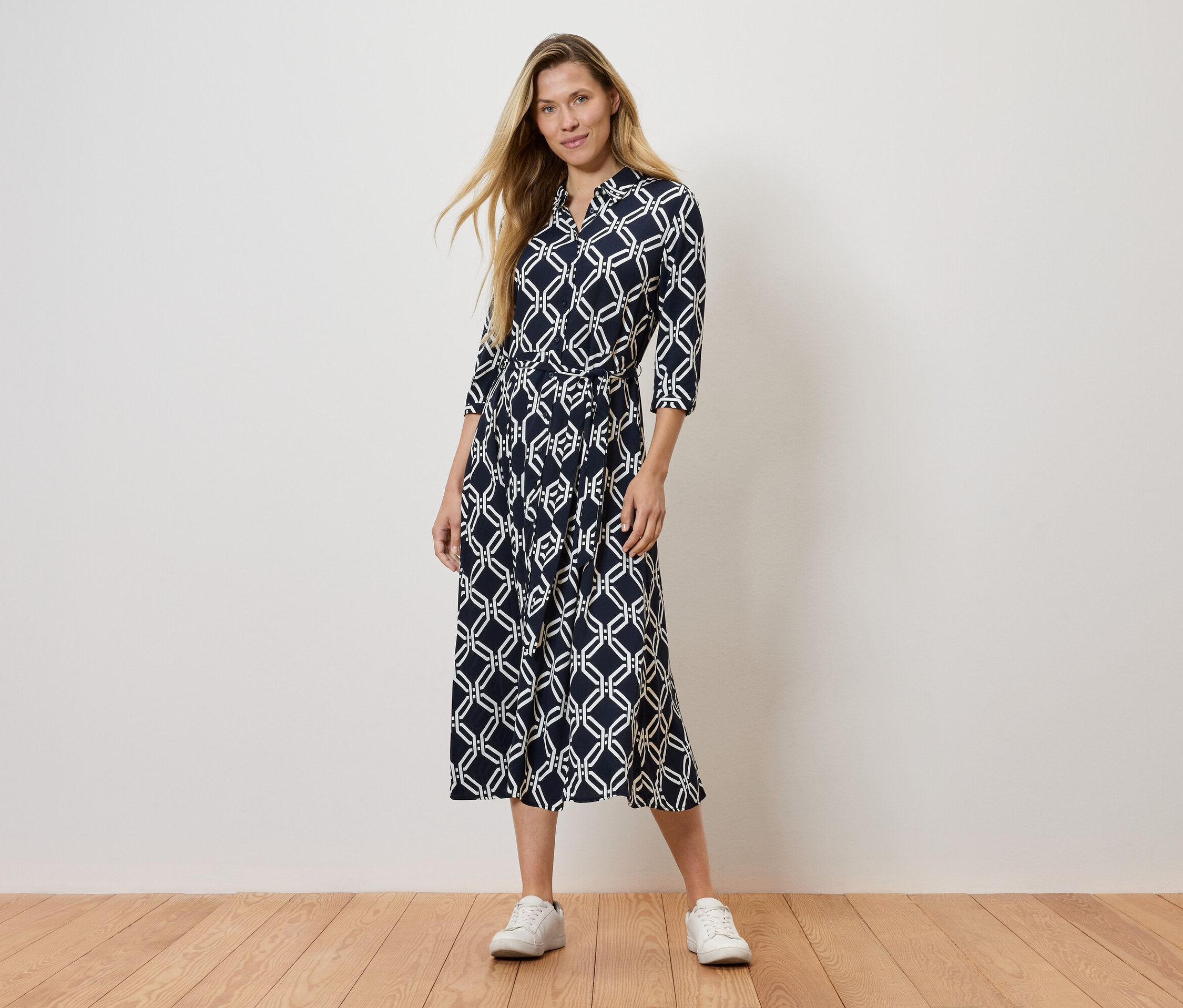 Kobieta stoi w granatowej sukience we wzory i białych tenisówkach.