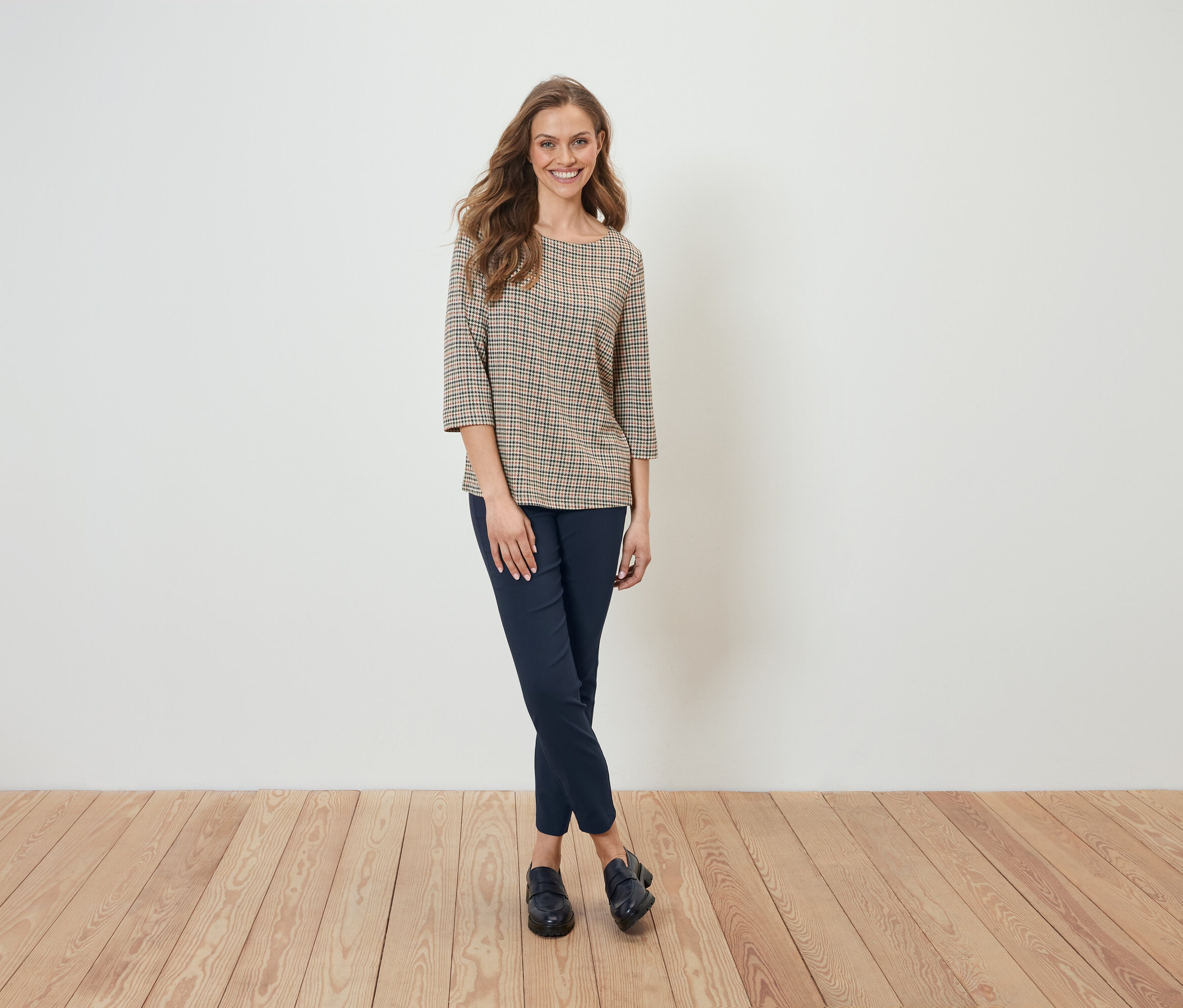 Kobieta w bluzce w kratę i granatowych spodniach stoi na tle białej ściany.