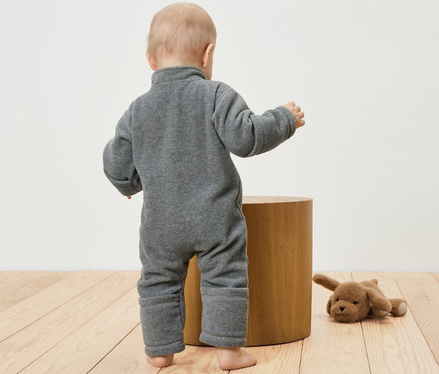 Niemowlę w szarym polarowym pajacyku stoi obok drewnianego stołka. Obok leży pluszowy pies.