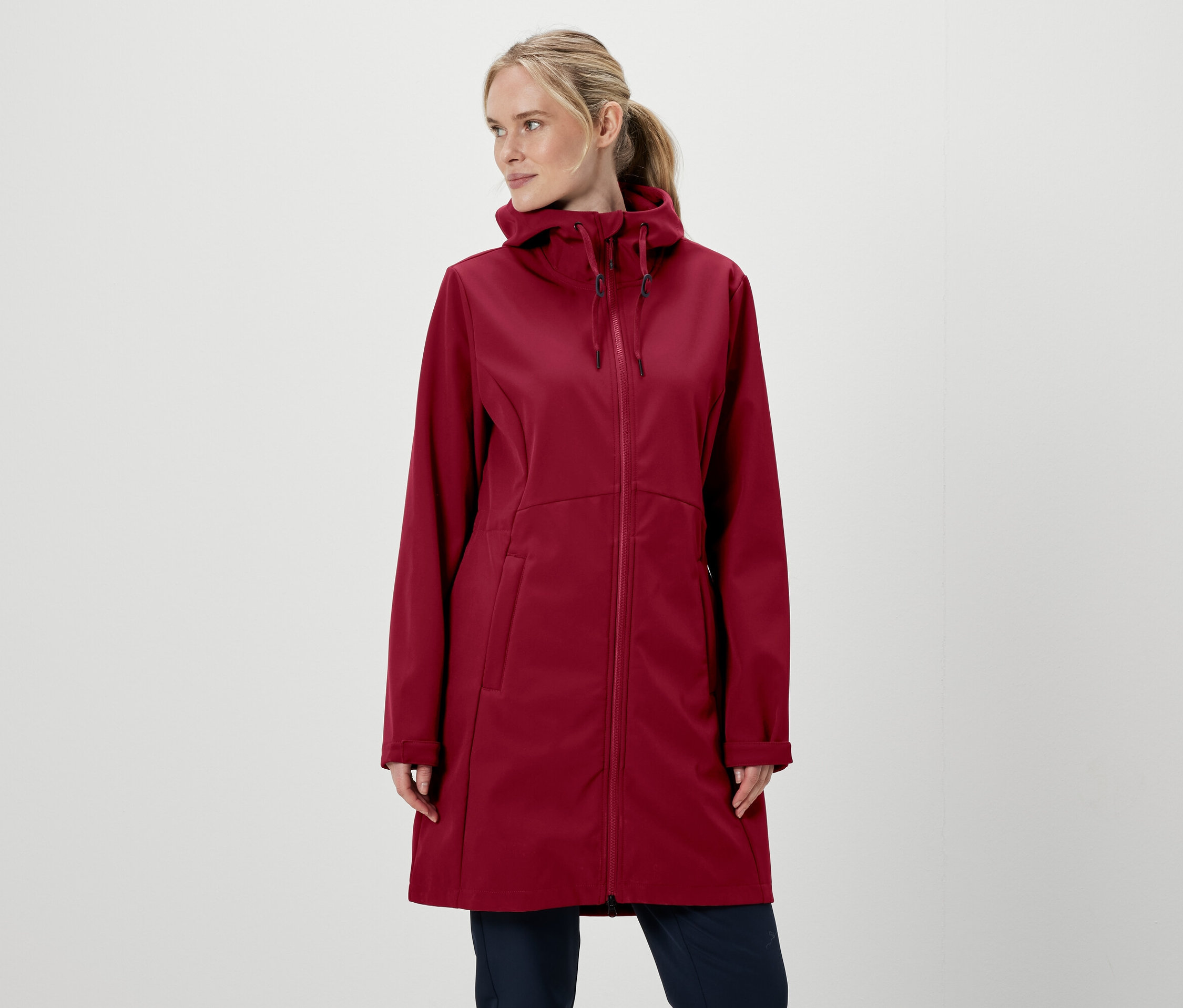 Blondynka w bordowej kurtce z kapturem zapinanej na zamek i spodniach na białym tle.