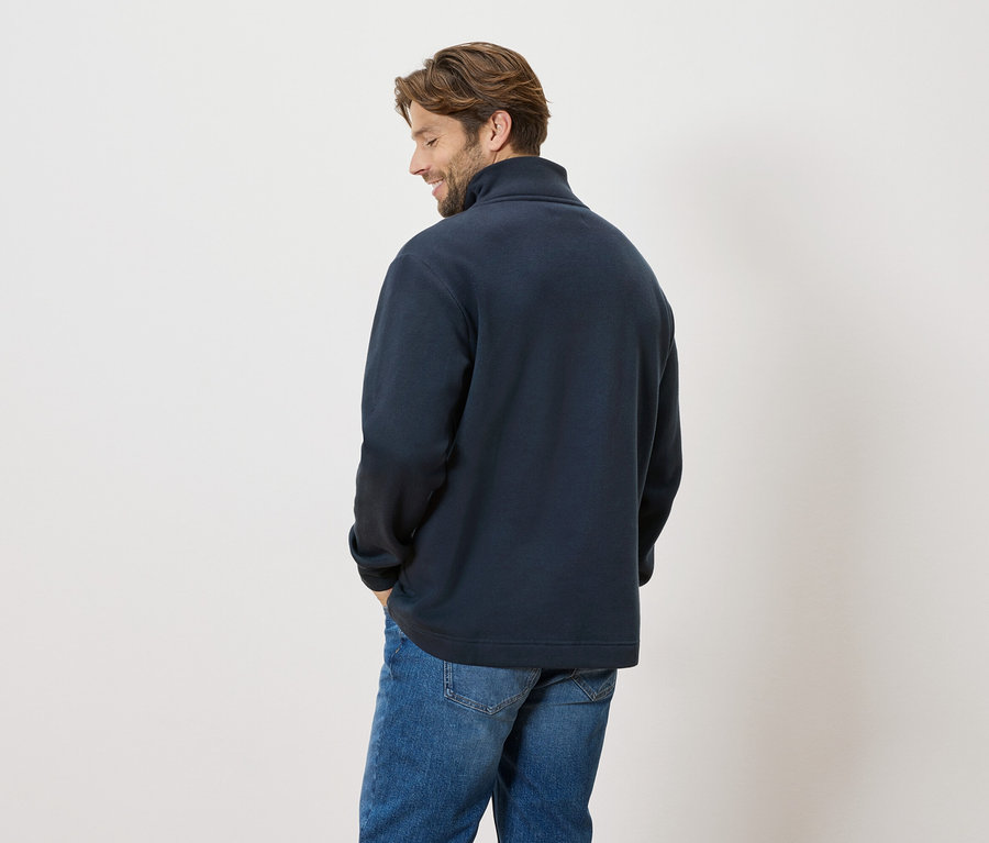 Mężczyzna stoi tyłem do kamery, ma na sobie granatowy sweter i dżinsy.