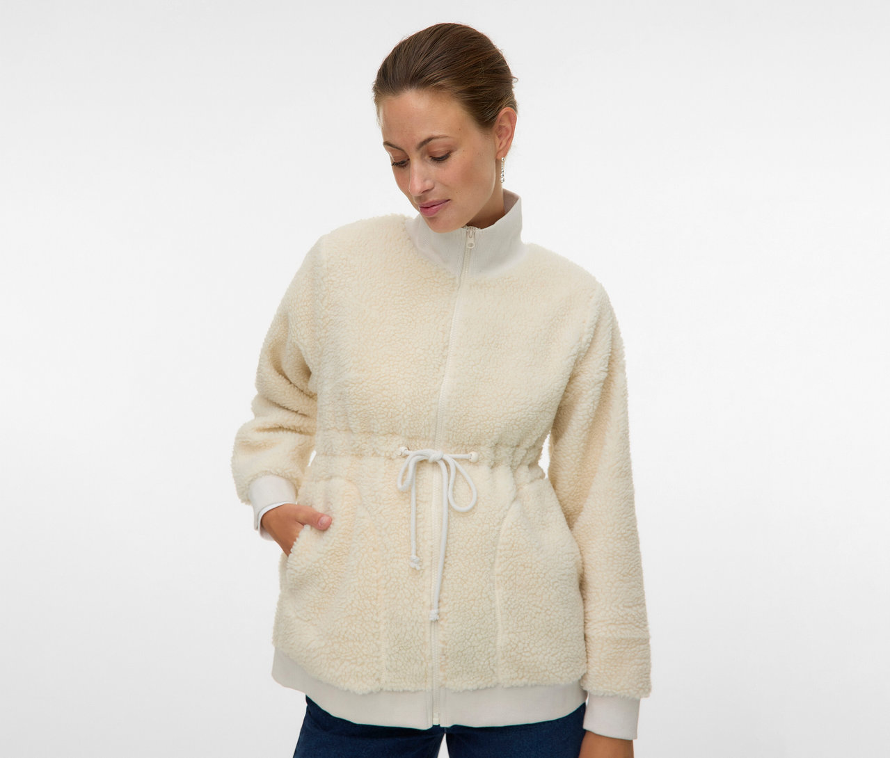 Kobieta ma na sobie rozpiętą, kremową bluzę pluszową 3 w 1 MAMALICIOUS „MLCARLY” ze sznurkiem w pasie i rękami w kieszeniach.