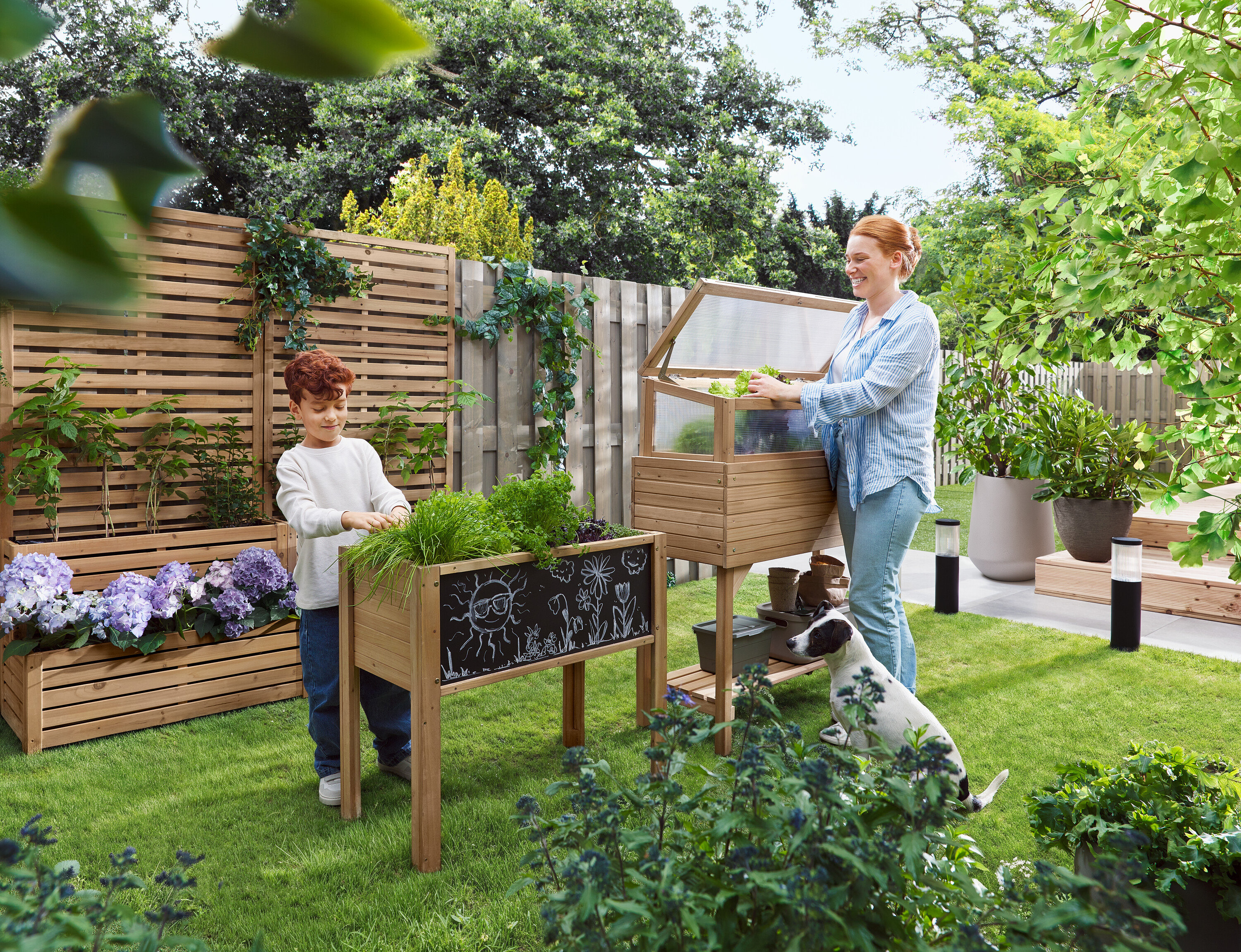Kobieta otwiera inspekt, podczas gdy dziecko stoi obok podwyższonej grządki. Pies siedzi na trawie.