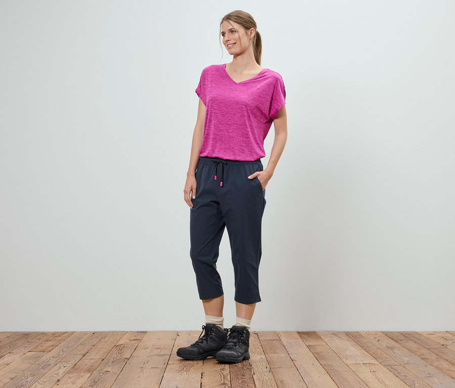 Kobieta stoi z rękami w kieszeniach na tle białej ściany. Ma na sobie różowy T-shirt, niebieskie spodnie trzy czwarte i czarne buty trekkingowe.