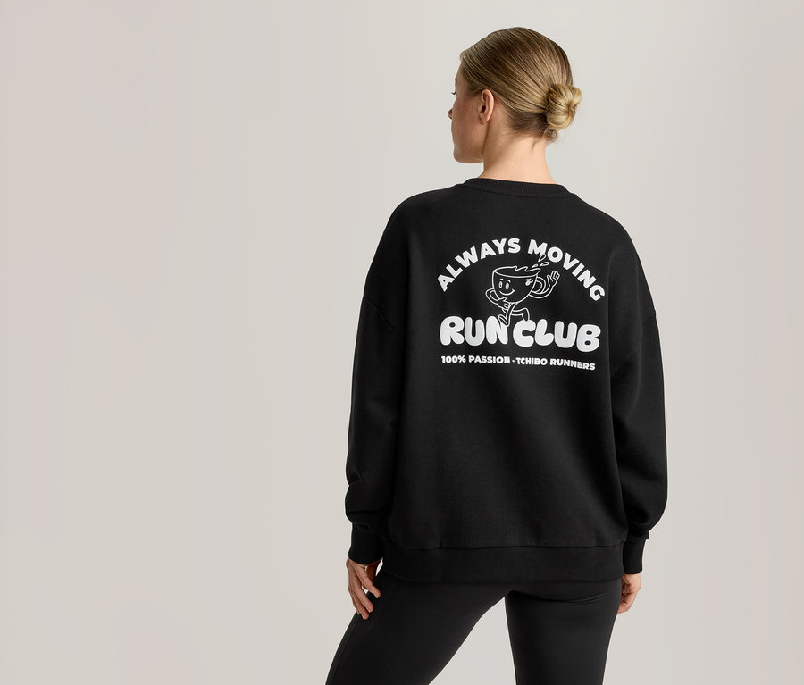 Kobieta widziana od tyłu nosi czarną bluzę z białym nadrukiem. Nadruk przedstawia kreskówkową filiżankę kawy oraz tekst 'ALWAYS MOVING RUN CLUB' i '100% PASSION - TCHIBO RUNNERS'.
