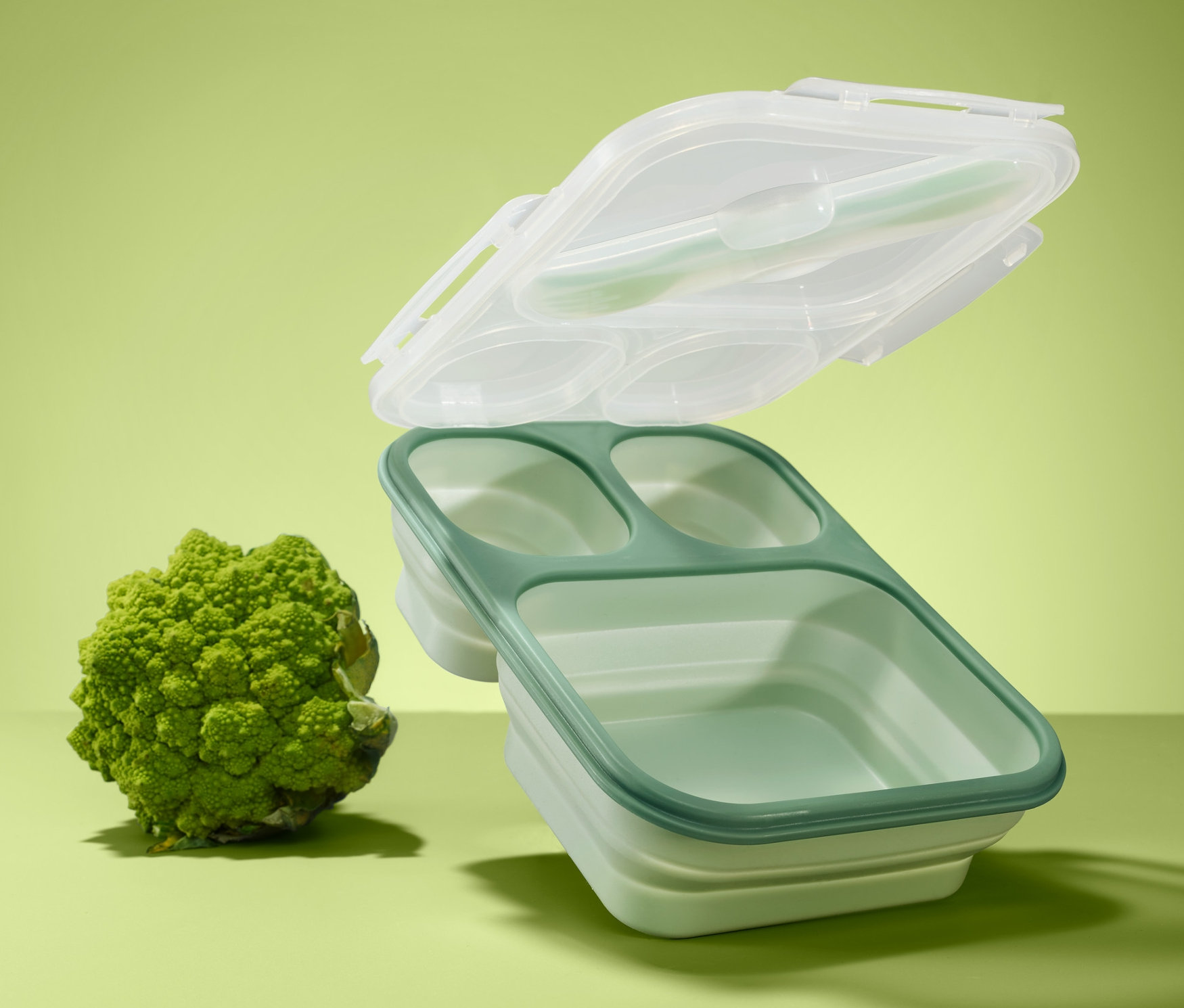 Zielony lunchbox składany z trzema przegródkami i przezroczystą pokrywką unosi się nad brokułem na zielonym tle.