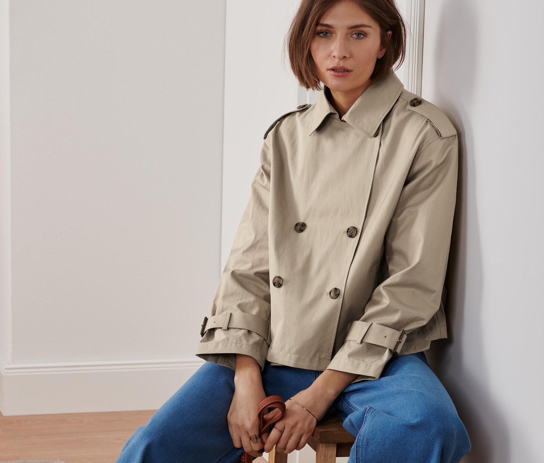 Kobieta siedzi na stołku w krótkim trenczu i trzyma brązową torebkę.