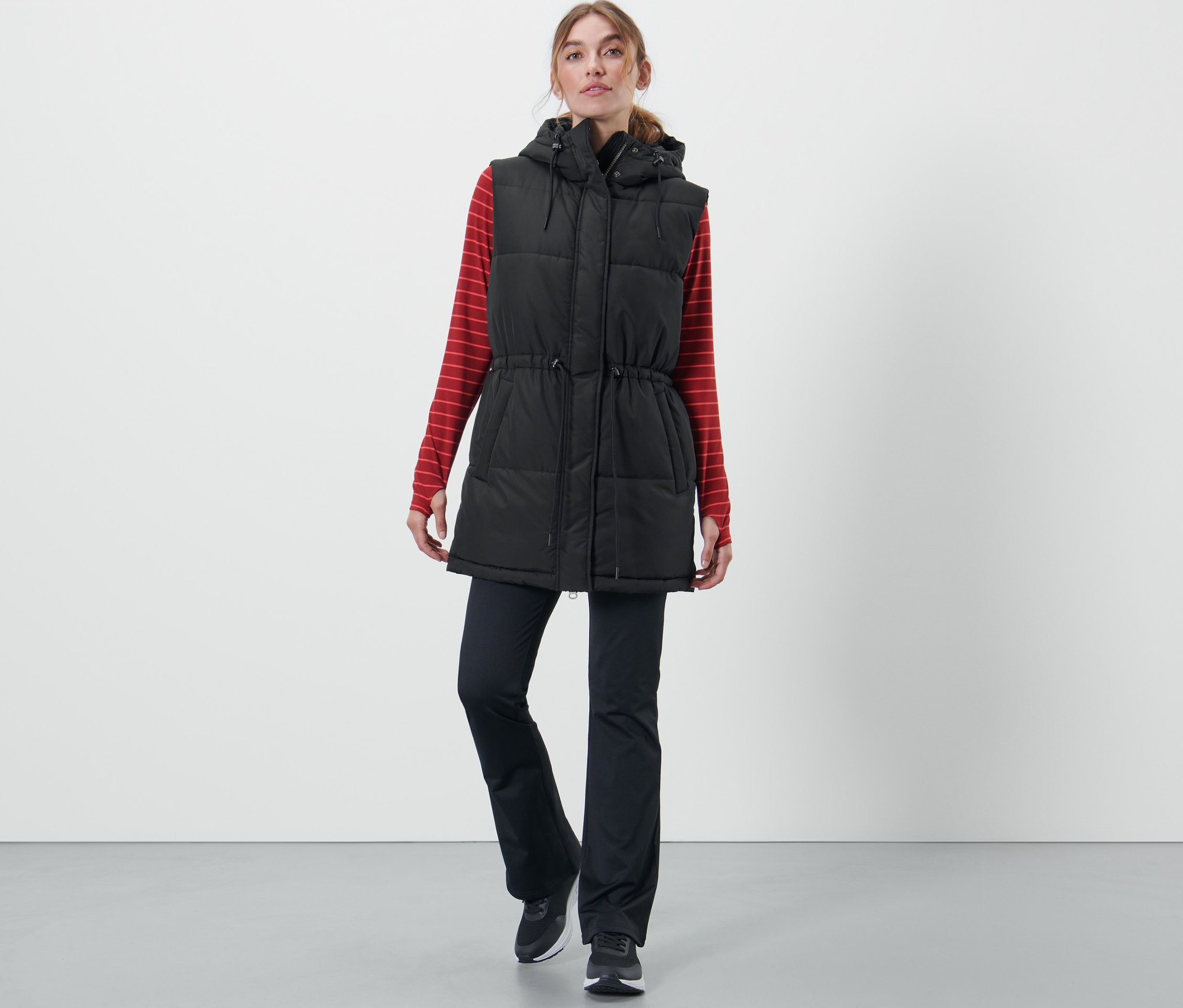 Kobieta ma na sobie pikowaną kamizelkę z kapturem, czerwoną bluzkę z długim rękawem w paski i czarne spodnie.