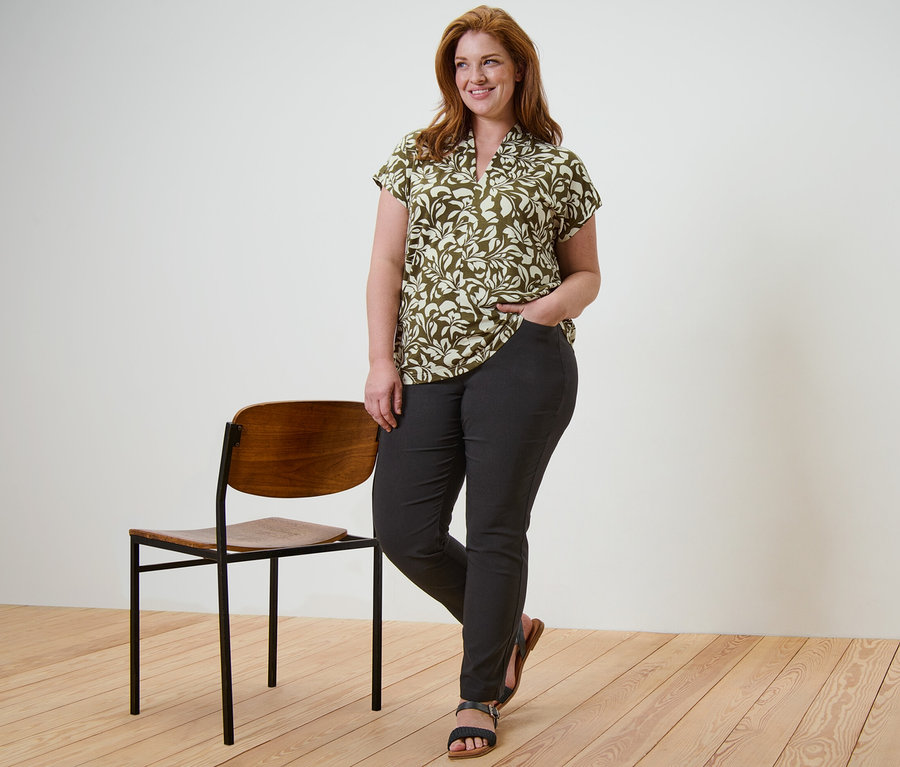 Kobieta stoi i pozuje w bluzce z dekoltem w serek i czarnych spodniach z bengaliny, z ręką w kieszeni, obok niej stoi krzesło.