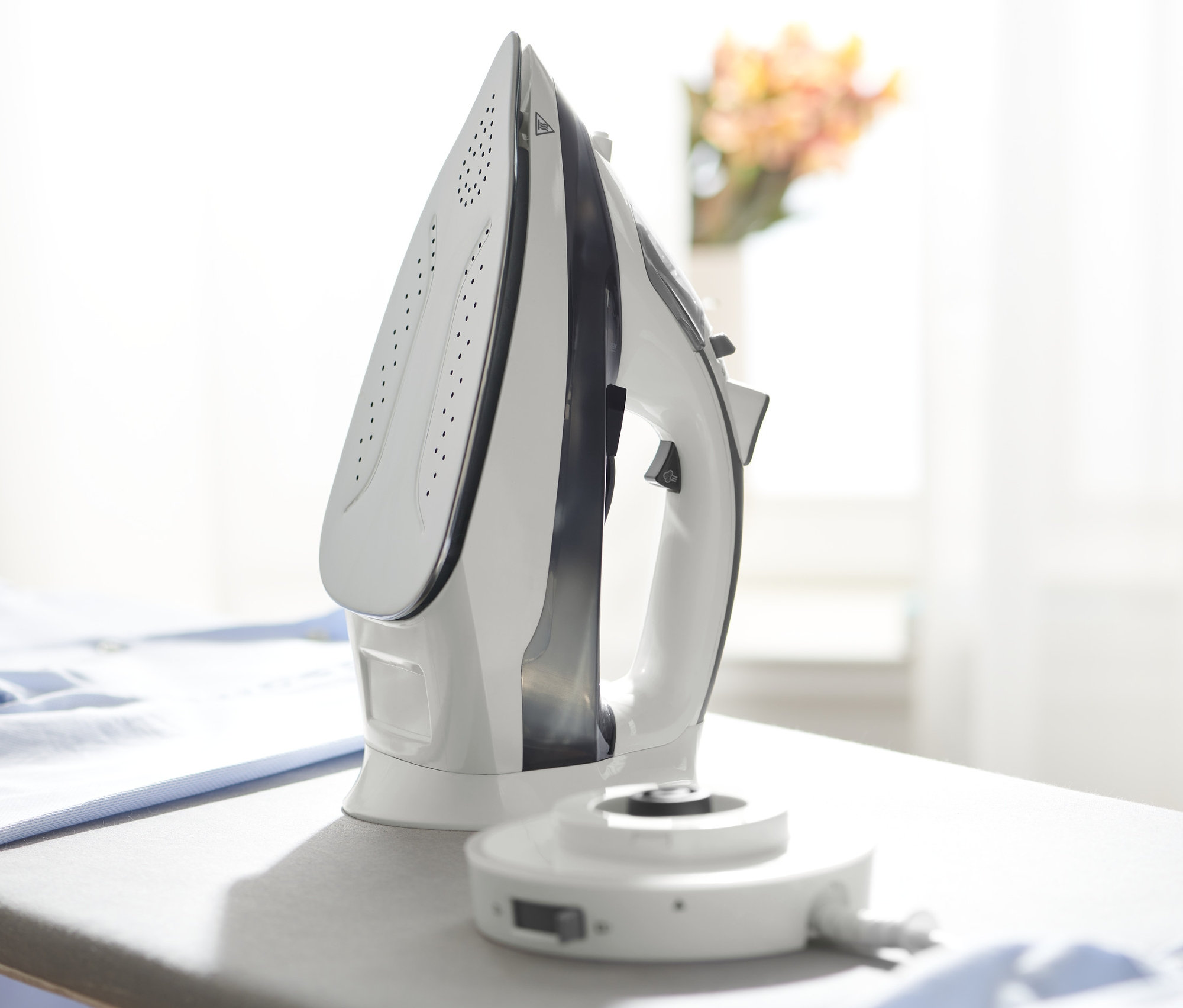 Białe bezprzewodowe żelazko parowe stoi na szarej desce do prasowania obok stosu niebieskich ubrań.