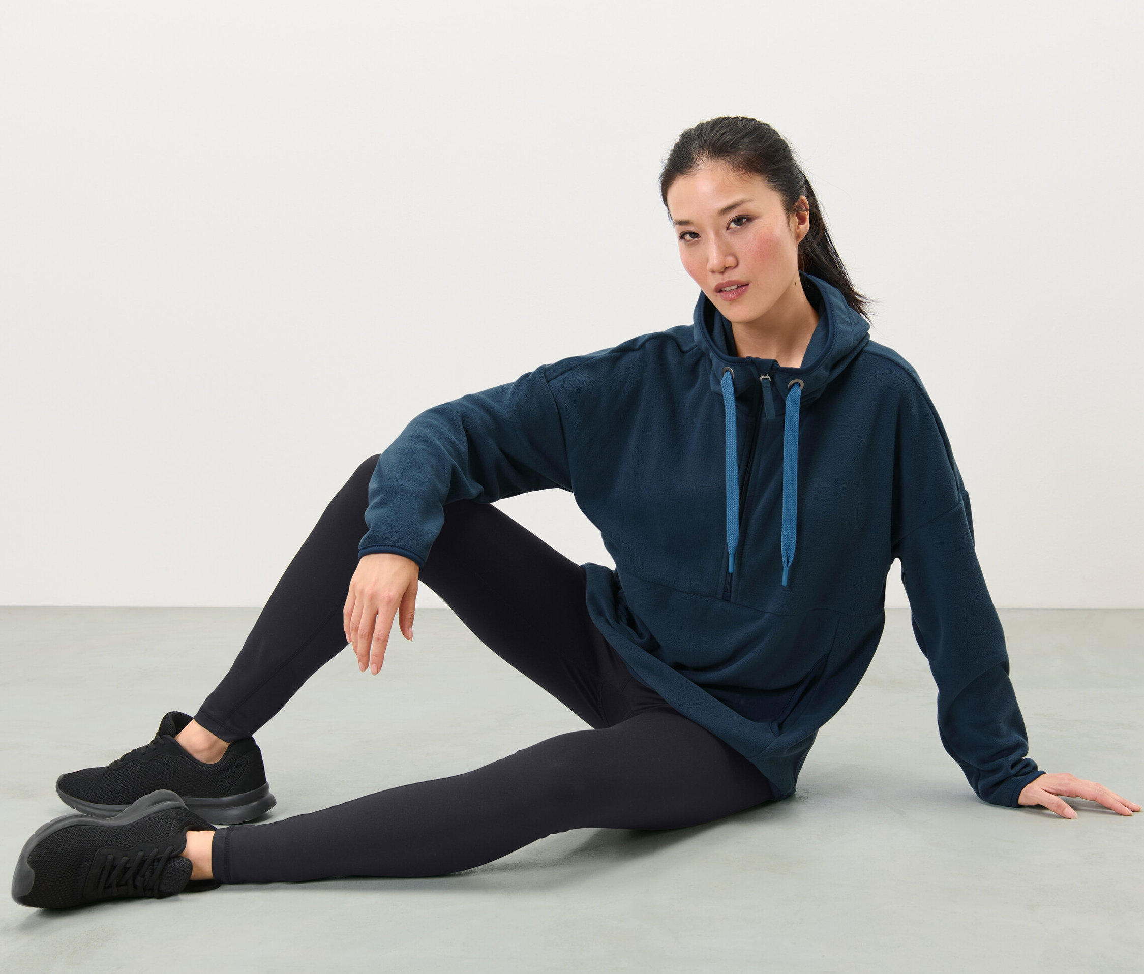 Kobieta siedzi na podłodze w polarowej bluzie z kapturem, czarnych legginsach i butach sportowych.