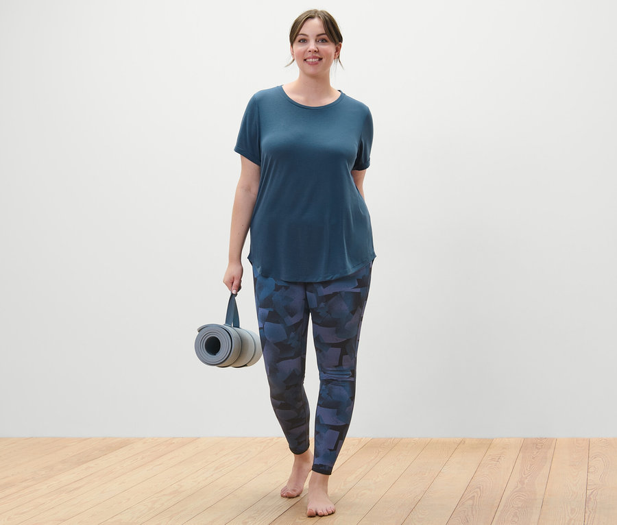 Kobieta w granatowej długiej koszulce i czarno-granatowych dwustronnych legginsach sportowych trzyma zwiniętą matę do ćwiczeń.