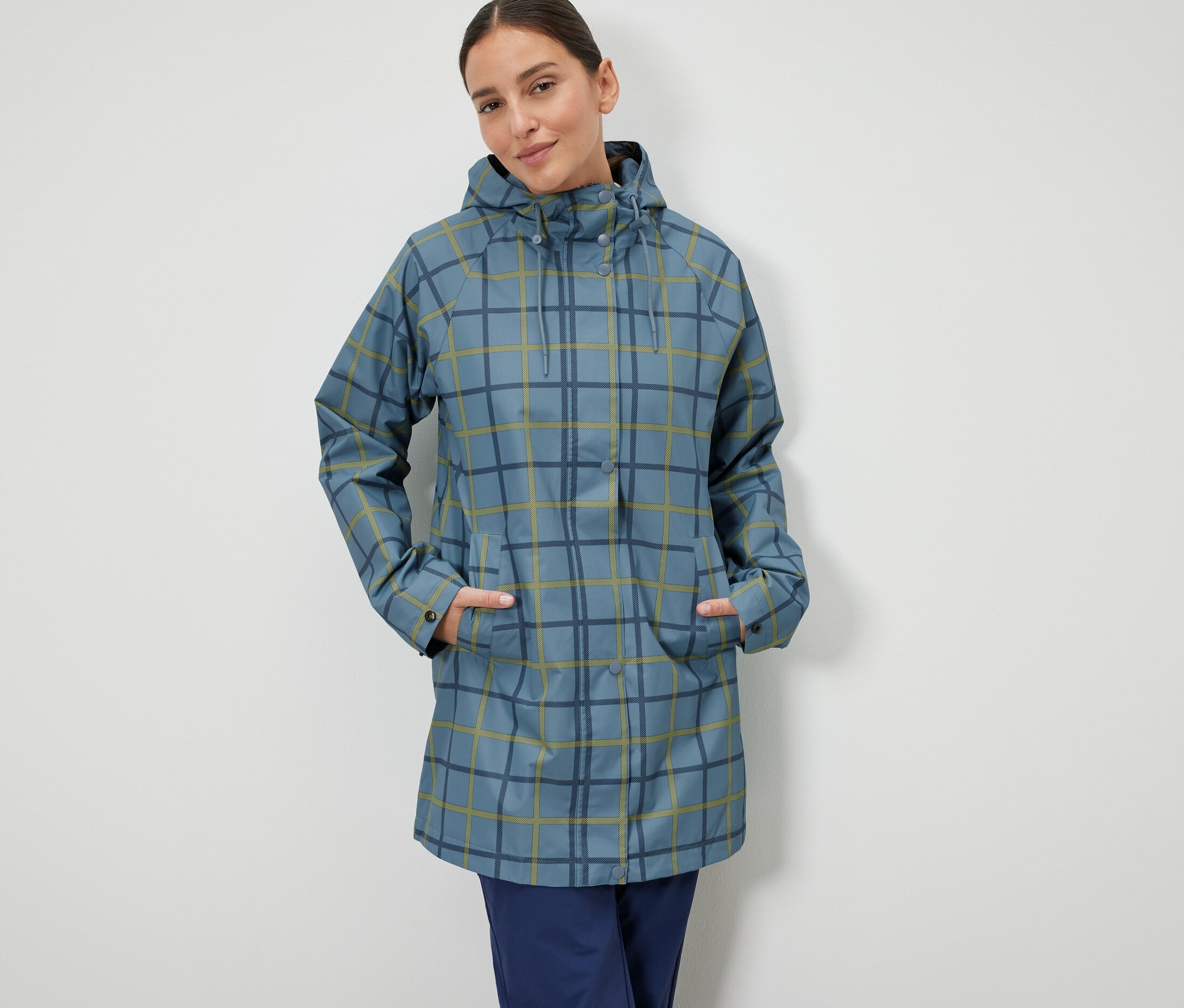 Kobieta stoi z rękami w kieszeniach niebieskiej kurtki przeciwdeszczowej w kratę.