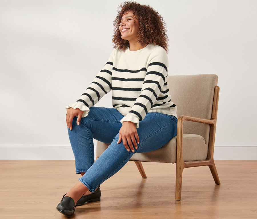 Kobieta siedzi w fotelu, ubrana w spodnie dżinsowe o kroju slimfit i sweter z falbanką w paski.