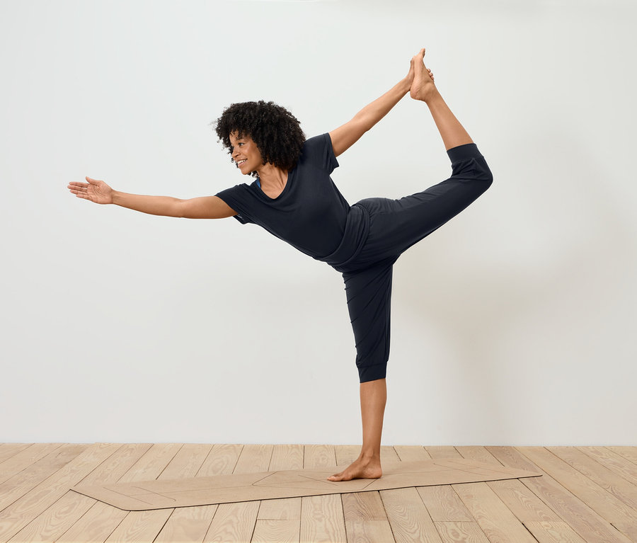 Kobieta w czarnym stroju sportowym stoi w pozycji tancerza na macie do jogi.