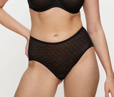 Osoba w czarnych figach Triumph Signature Sheer Maxi EX z wzorzystego materiału i pasującym biustonoszu, z rękami na biodrach.