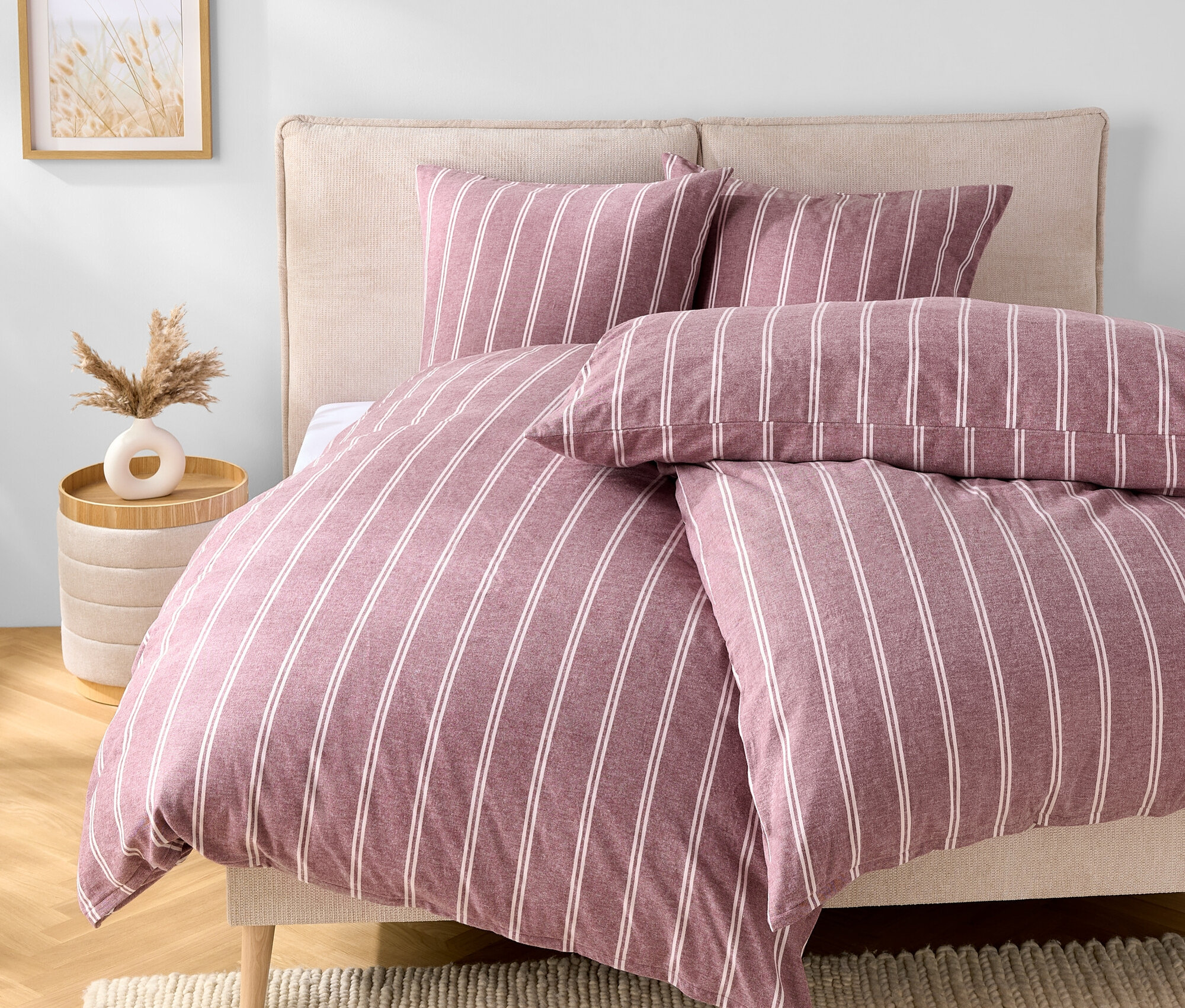 Sypialnia z łóżkiem z fioletowo-białą pościelą w paski, dwiema poduszkami i beżowym zagłówkiem. Obok łóżka stoi stolik z wazonem.