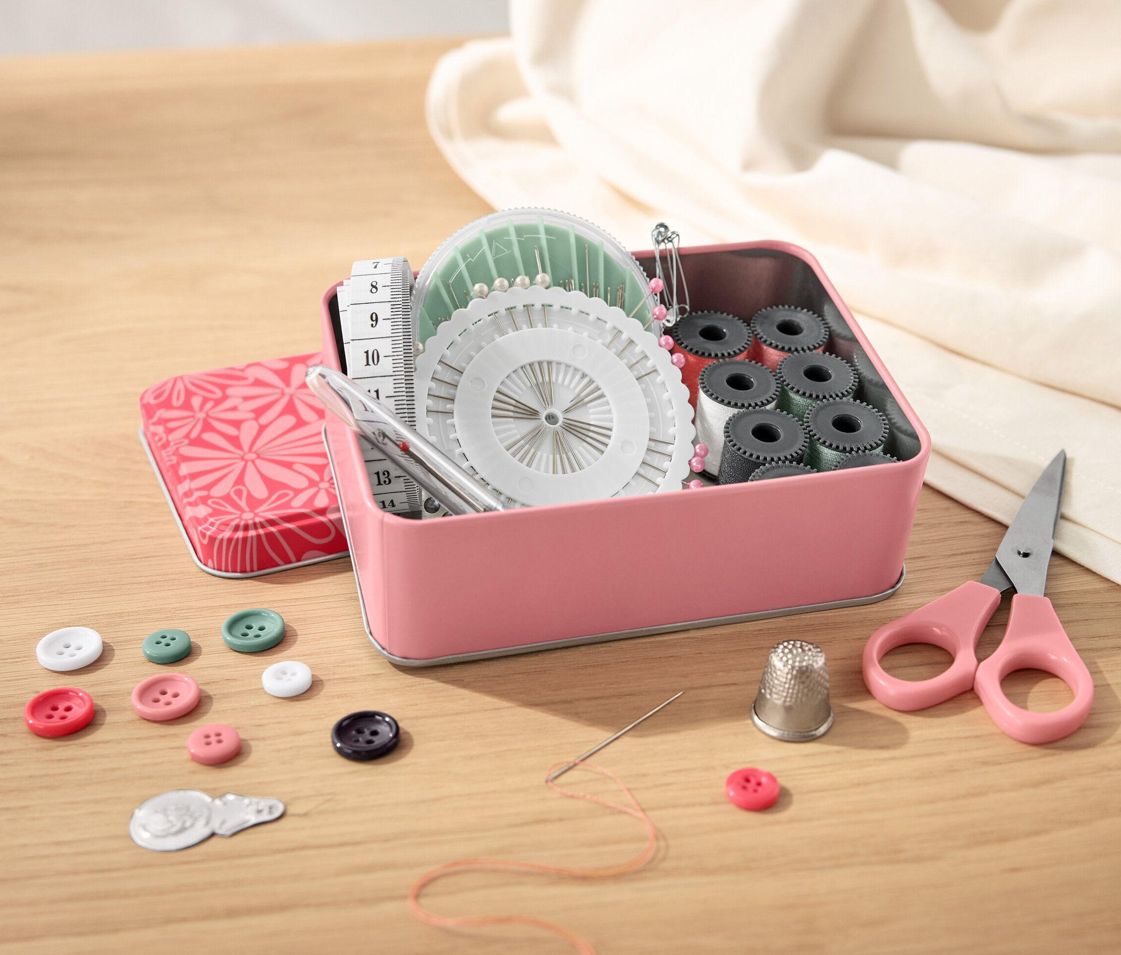 Otwarty metalowy pojemnik z zestawem do szycia, guzikami, nożyczkami i miarką krawiecką na drewnianym stole.