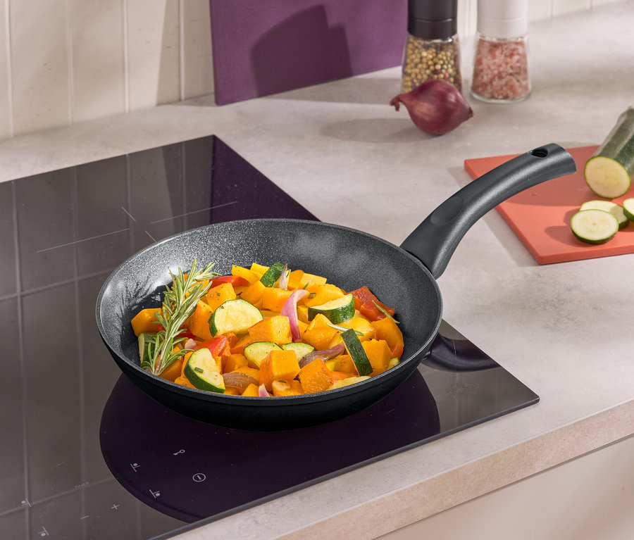 Patelnia z smażonymi warzywami na płycie indukcyjnej. Obok patelni leży pokrojona cukinia na desce.