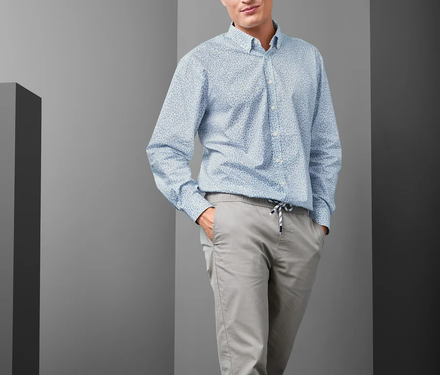 Mężczyzna pozuje, ubrany w jasnoniebieską koszulę z kołnierzykiem button down Comfort Fit, szare spodnie i niebieskie tenisówki.