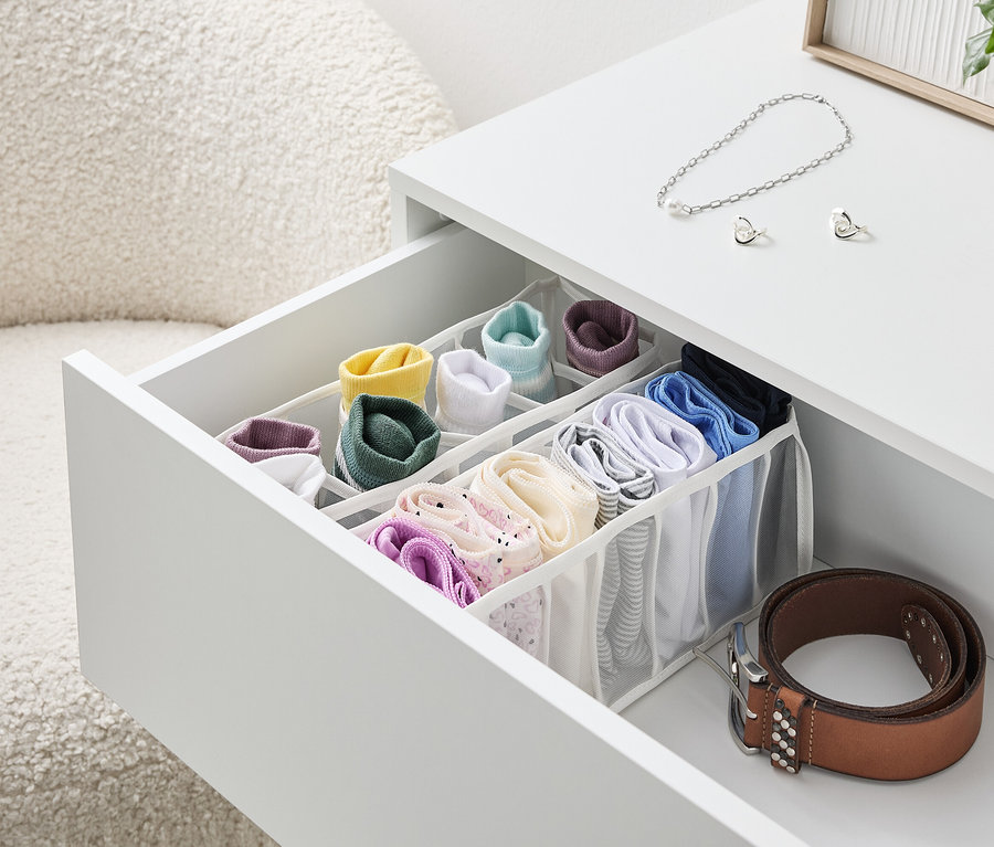 Otwarta szuflada białego stolika z organizerem pełnym kolorowych skarpetek i bielizny, obok leży brązowy pasek.