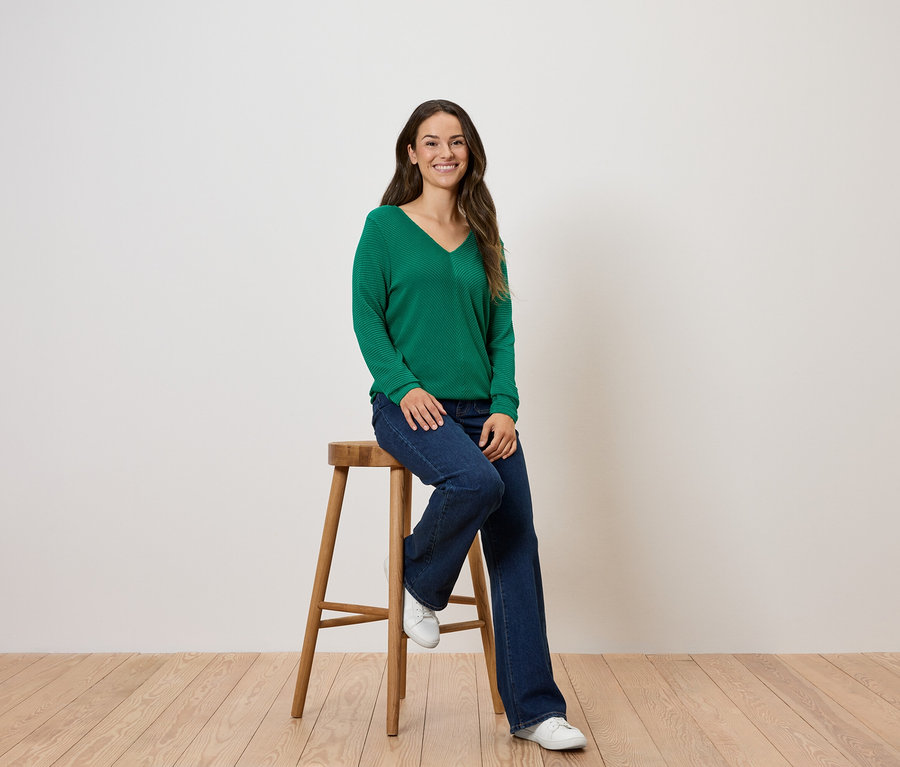 Kobieta siedzi na drewnianym stołku, ubrana w zielony sweter i dżinsy.