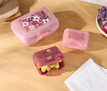 Trzy kolorowe pudełka na lunch z różnymi wzorami, chleb i owoce na drewnianym stole.