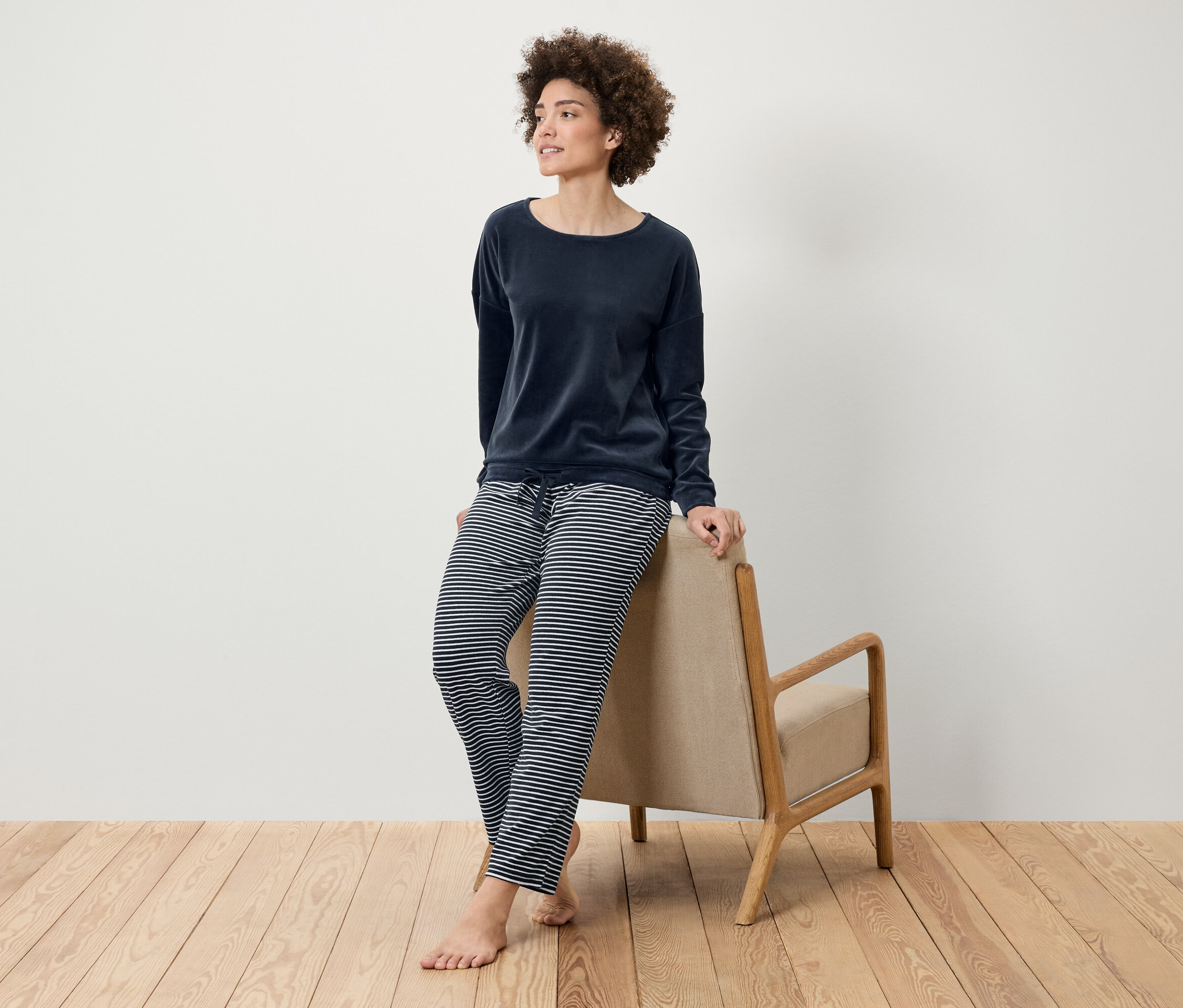 Kobieta z fryzurą afro pozuje w granatowym welurowym topie i spodniach w paski, opierając się o beżowy fotel na drewnianej podłodze.