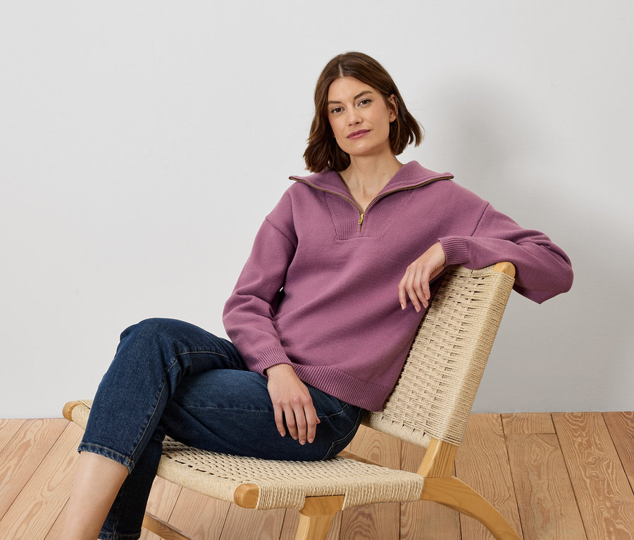 Kobieta siedzi na krześle, ubrana w fioletowy sweter i dżinsy.