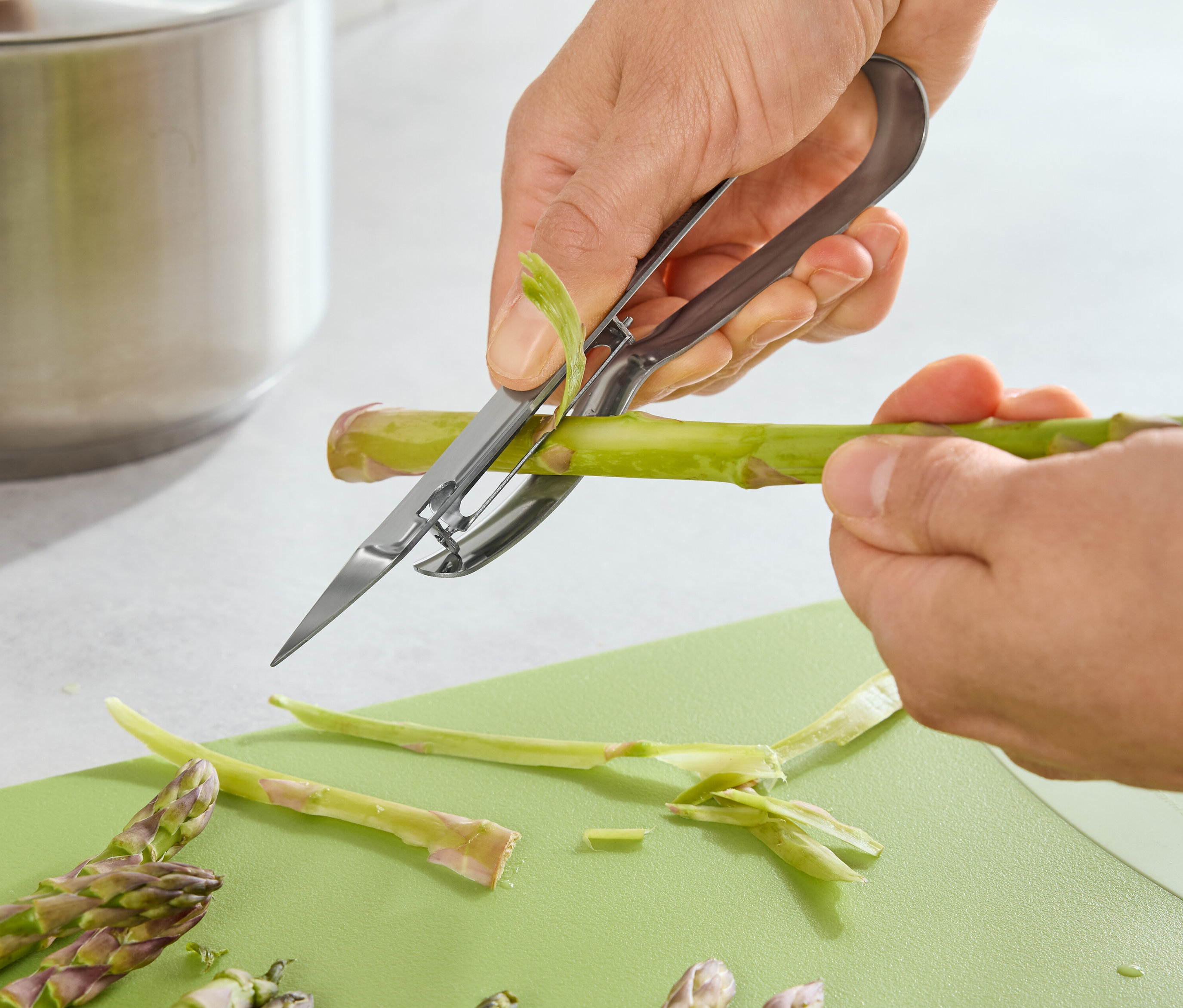 Osoba obiera szparagi obieraczką do szparagów na zielonej desce do krojenia.