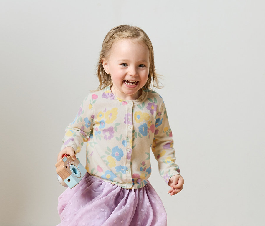 Uśmiechnięta blondwłosa dziewczynka z zaplecionymi warkoczami w kwiecistym kardiganie i fioletowej spódnicy trzyma zabawkę-aparat.