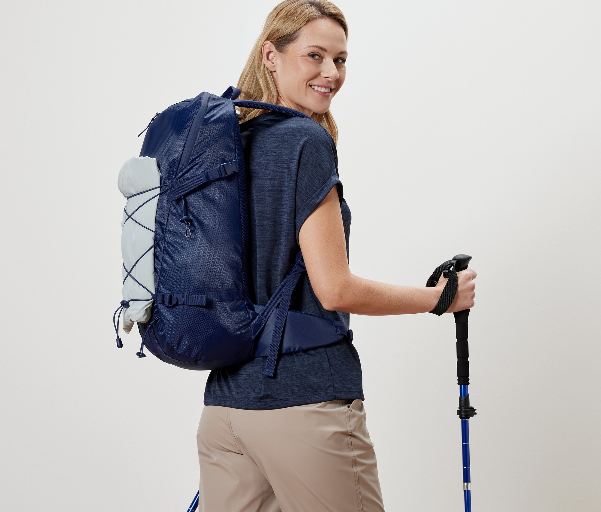 Kobieta z plecakiem turystycznym i kijkami trekkingowymi spogląda przez ramię.