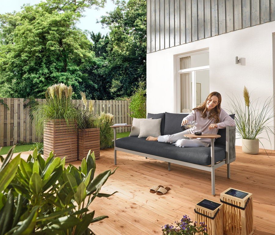 Kobieta nalewa wodę do szklanki na tarasie. Obok niej stoi bambusowa latarnia solarna, mała i duża, 5 sztuk solarnych lamp LED na szpikulcach i sofa ogrodowa z 2 stolikami obrotowymi oraz 2 donice-kolumny na rośliny »Tinus«.