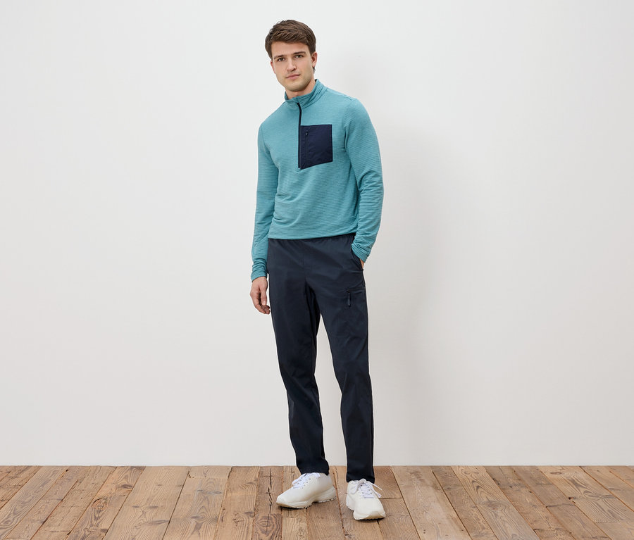 Mężczyzna stoi na tle białej ściany, ubrany w turkusową bluzę z długim rękawem z ciemną kieszenią, granatowe spodnie i białe tenisówki.