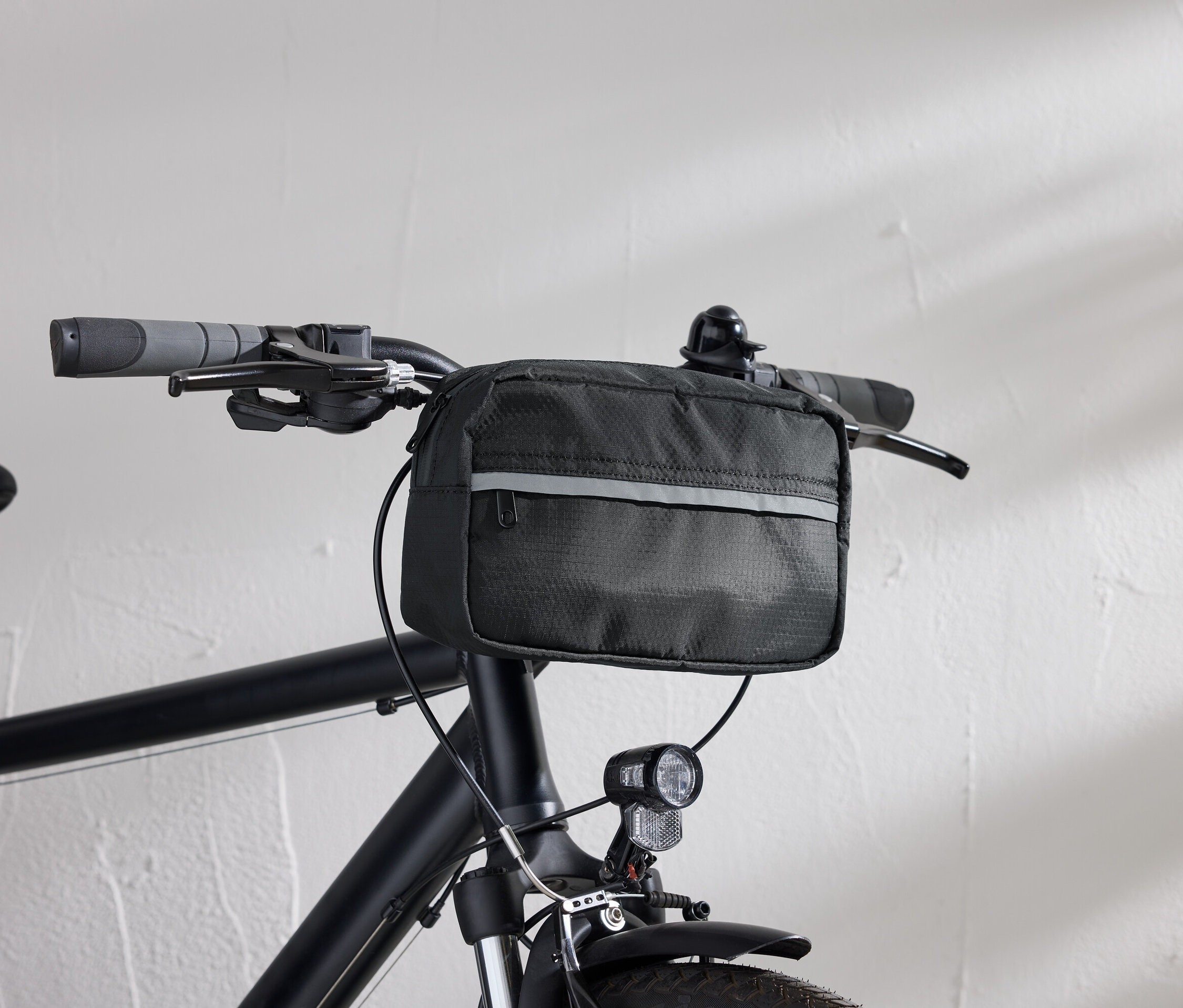 Szczegółowy widok czarnego roweru z czarną torbą na kierownicę i lampką rowerową na tle białej ściany.