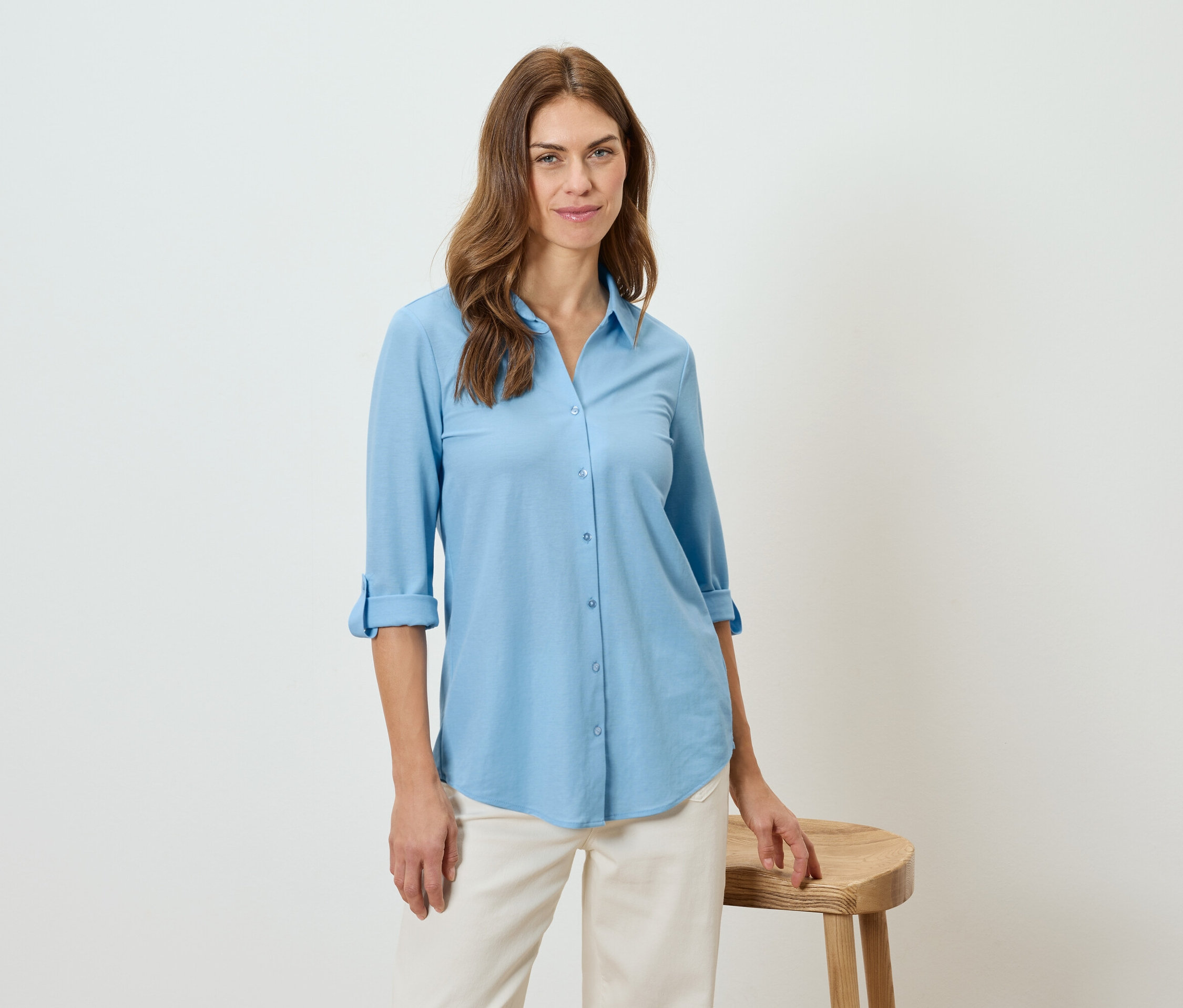 Kobieta ma na sobie błękitną koszulę i beżowe spodnie, opiera się o stołek.