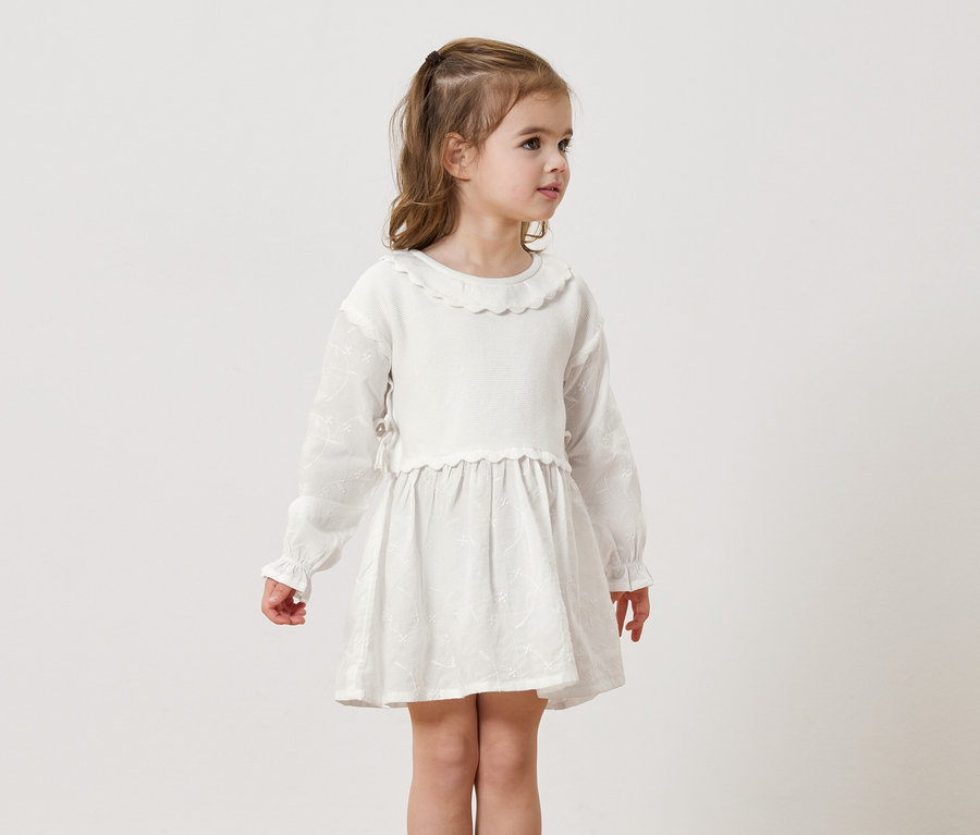 Dziewczynka z blond włosami ma na sobie białą sukienkę z długimi rękawami na neutralnym tle.