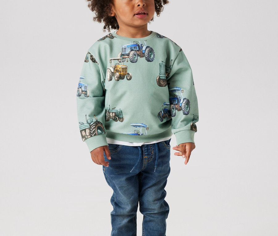 Mały chłopiec z kręconymi włosami stoi w miętowo-zielonym swetrze z traktorami i dżinsach.