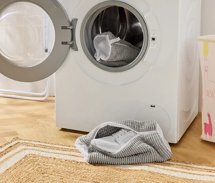 Szara torba na buty z białym tenisówką stoi przed pralką z otwartymi drzwiami. Kolejna torba na buty znajduje się w bębnie.
