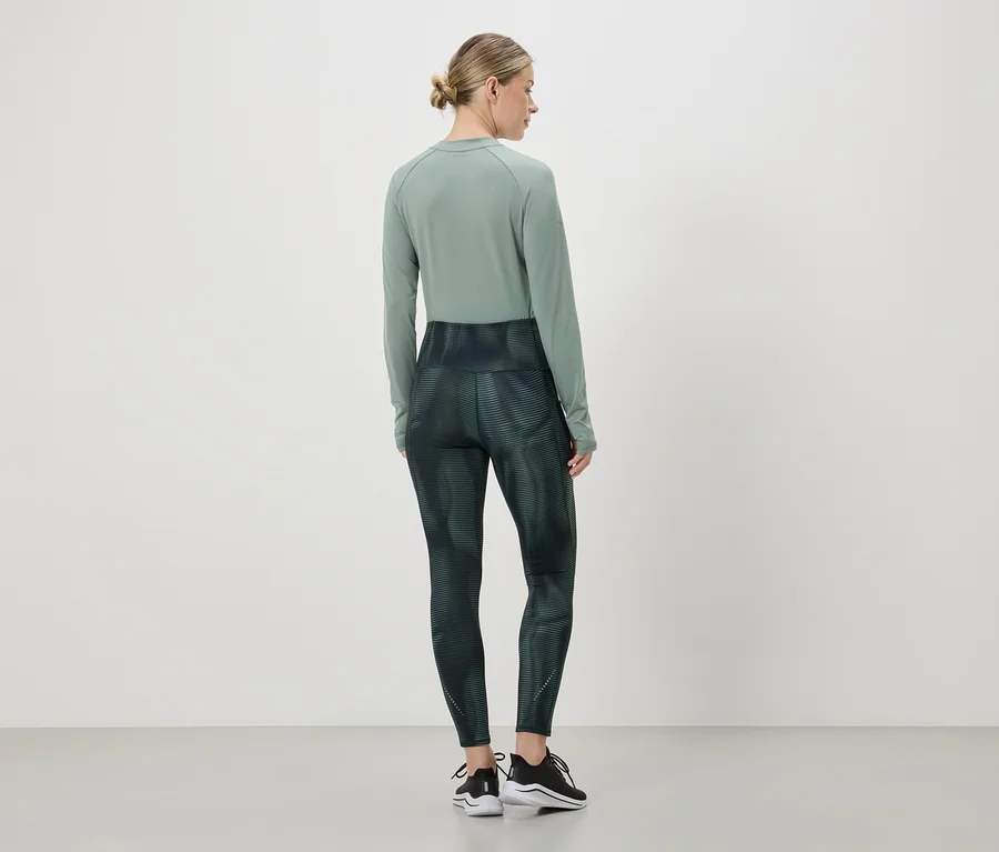 Kobieta z kokiem ma na sobie zieloną koszulę z długim rękawem i legginsy z abstrakcyjnym wzorem oraz czarne buty sportowe.