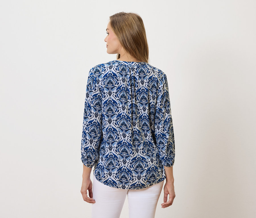 Kobieta z długimi brązowymi włosami ma na sobie bluzkę w niebiesko-białe wzory i białe spodnie.