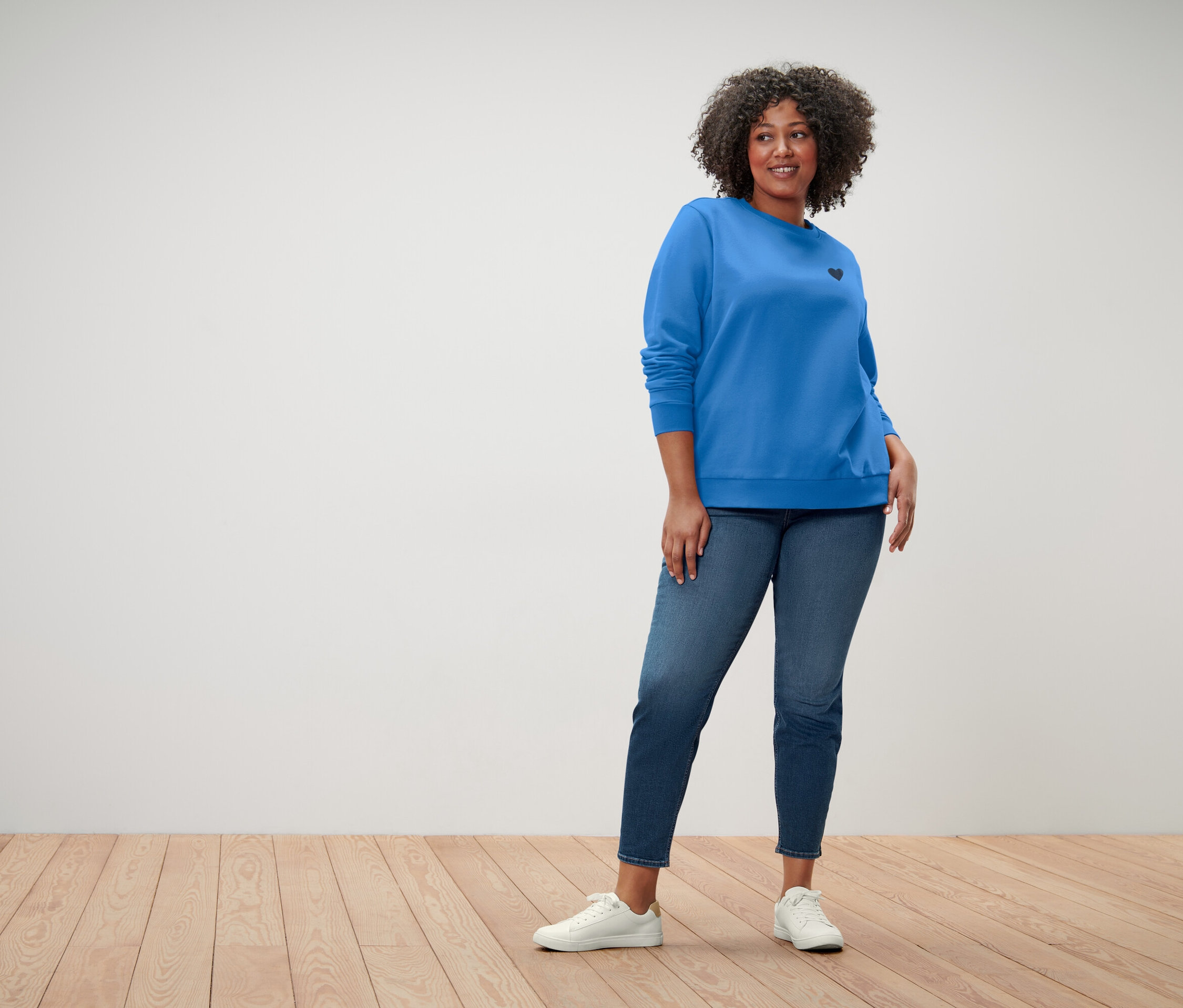 Kobieta z kręconymi włosami ma na sobie niebieską bluzę z małym czarnym sercem, niebieskie dżinsy i białe trampki.