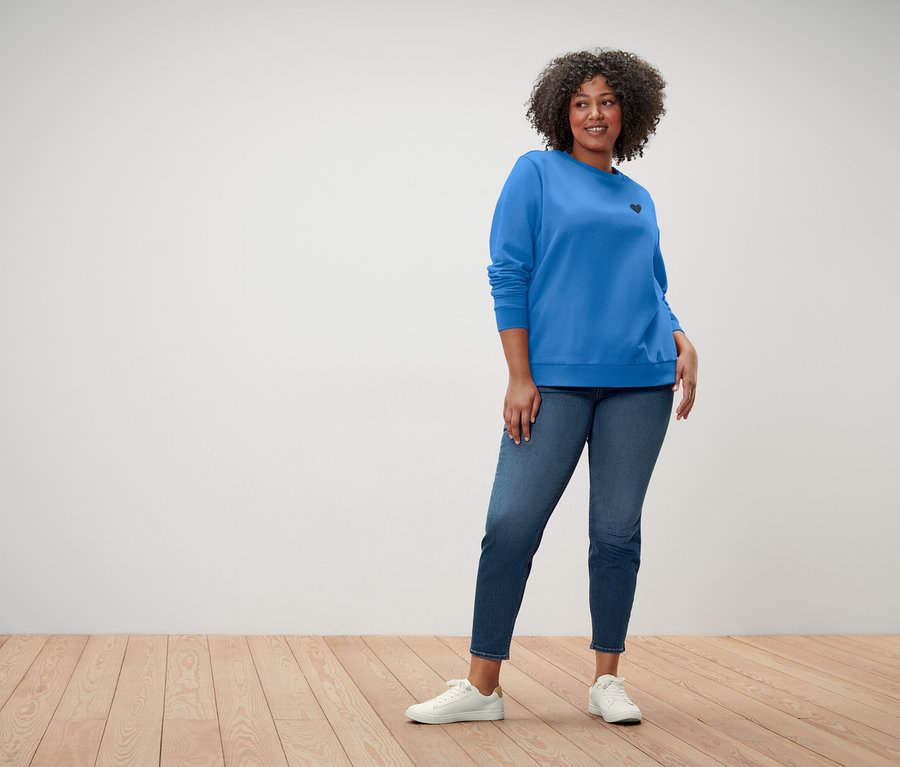 Kobieta z kręconymi włosami ma na sobie niebieską bluzę z małym czarnym sercem, niebieskie dżinsy i białe trampki.