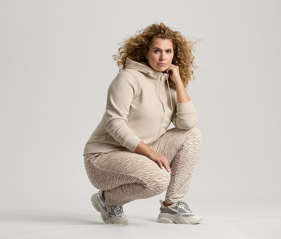 Kobieta z kręconymi włosami w beżowej kurtce, spodniach w zebrę i butach sportowych kuca z ręką na brodzie.