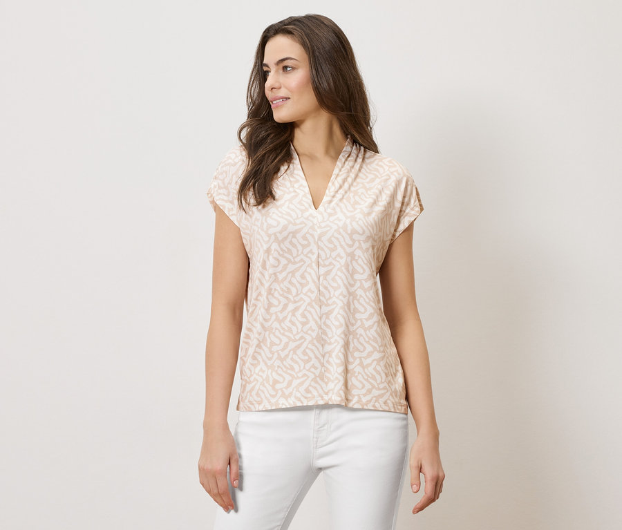 Kobieta ma na sobie jasno brązową bluzkę we wzory z dekoltem w serek i białe spodnie.